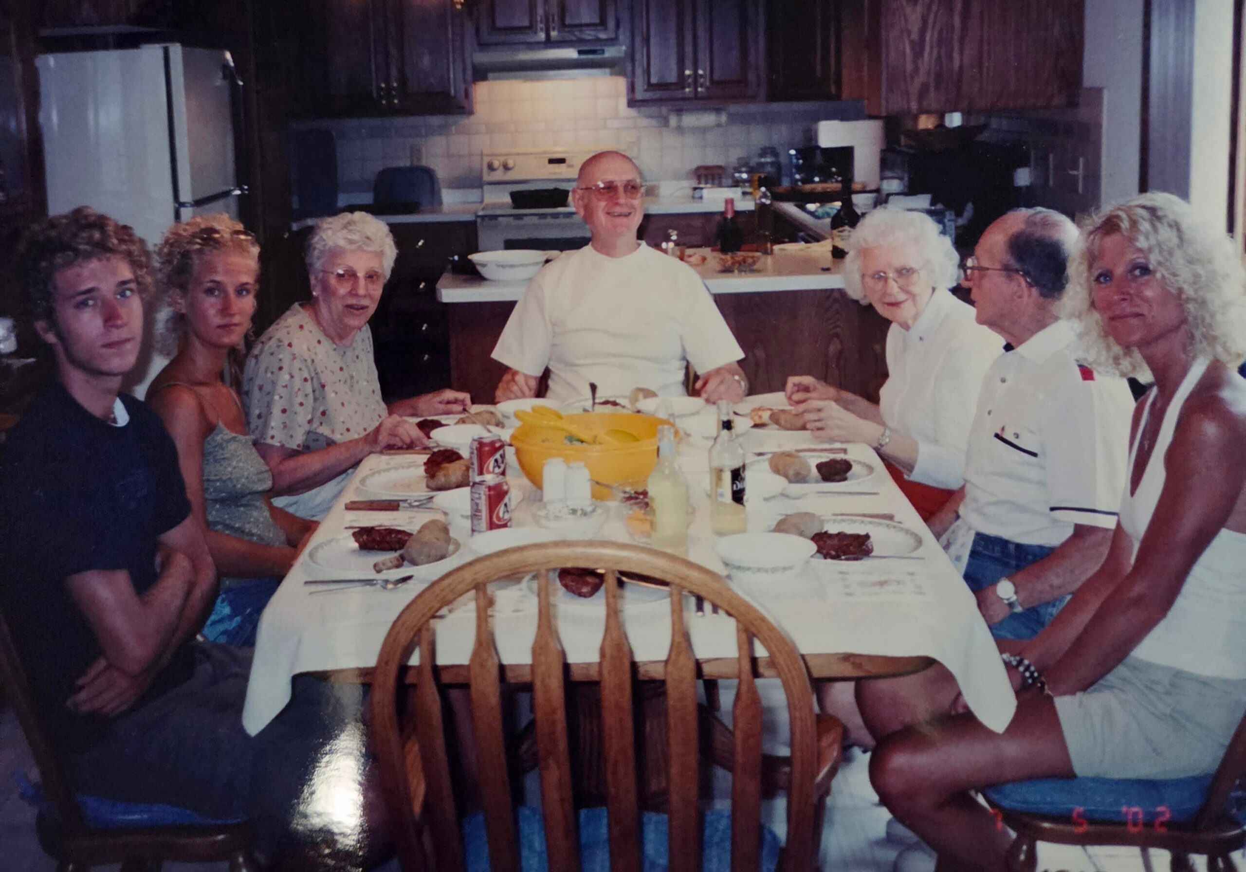 A family of seven seated around a kitchen table, sharing a meal with bread, drinks, and a yellow fruit bowl in the center of the table.