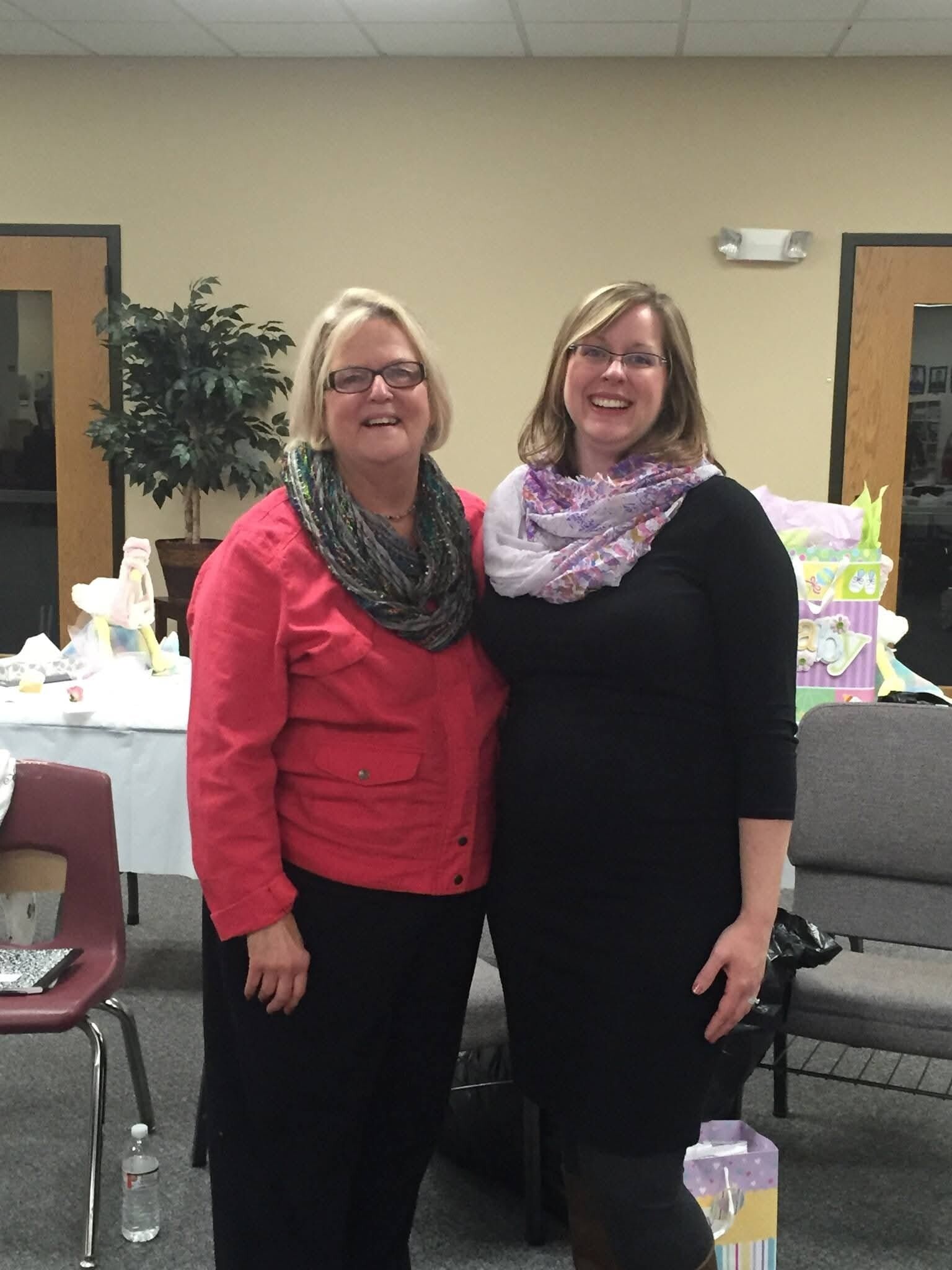 Two smiling women posing together indoors at an event, with gift bags and tables in the background.
