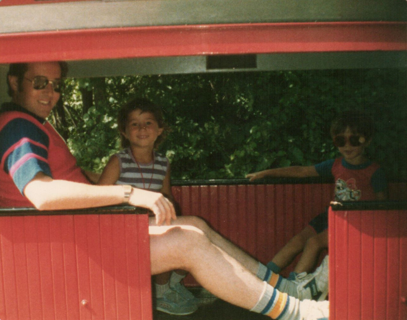 Man wearing sunglasses sits in a red wooden train car with two smiling children.