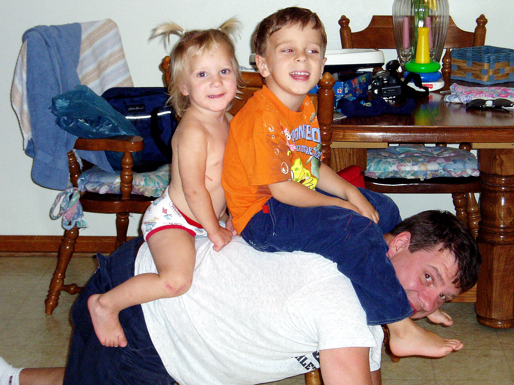 Man lying on a sofa with two young children climbing over him playfully in a living room scene.