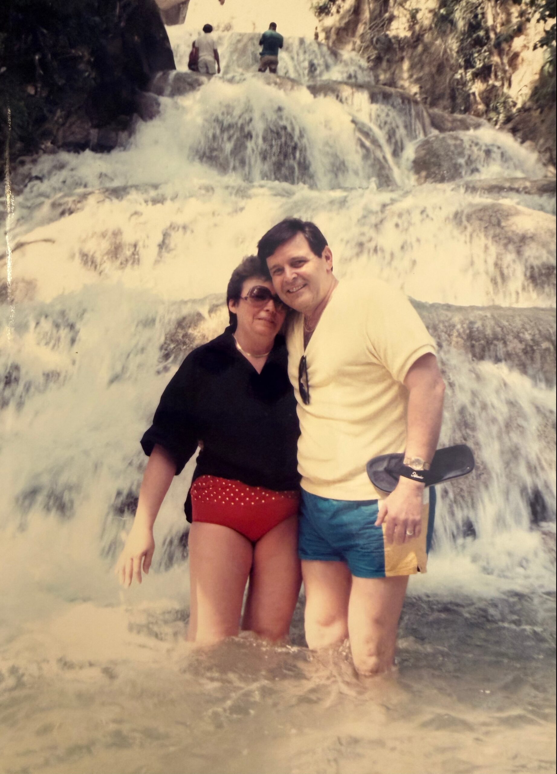 A couple posing together in swimwear at a cascading waterfall, water splashing around their legs.