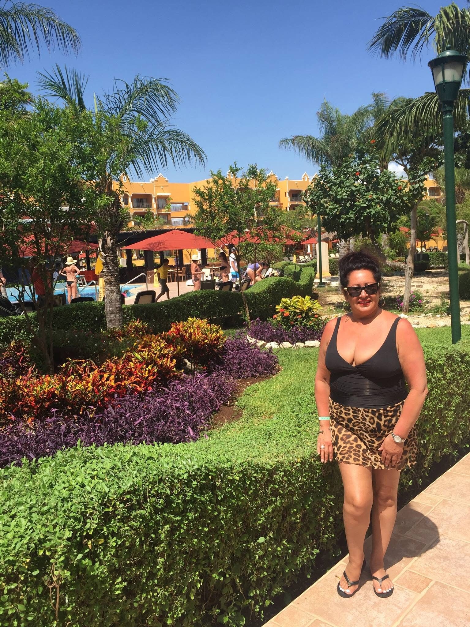 Woman in a black swimsuit and leopard print skirt stands on a sunny resort path beside colorful landscaping and hedges.