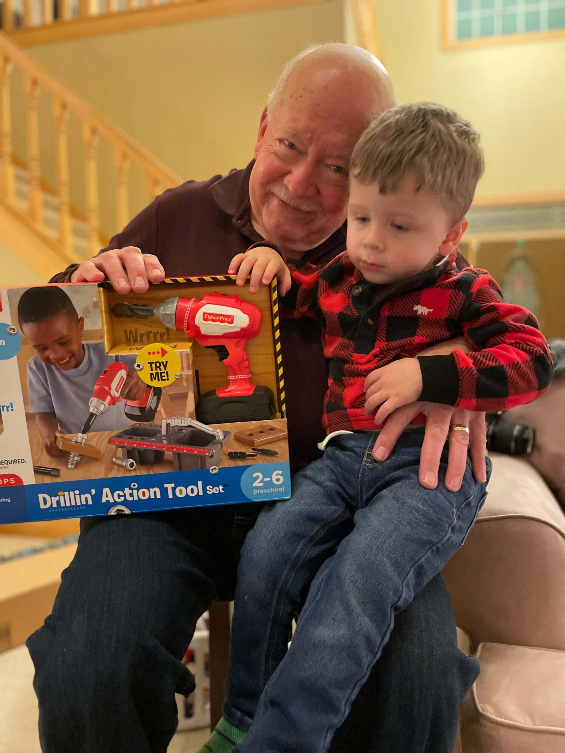 Grandfather and young boy smile as they hold a Drillin' Action Tool Set box together indoors, showcasing a toy drill kit.