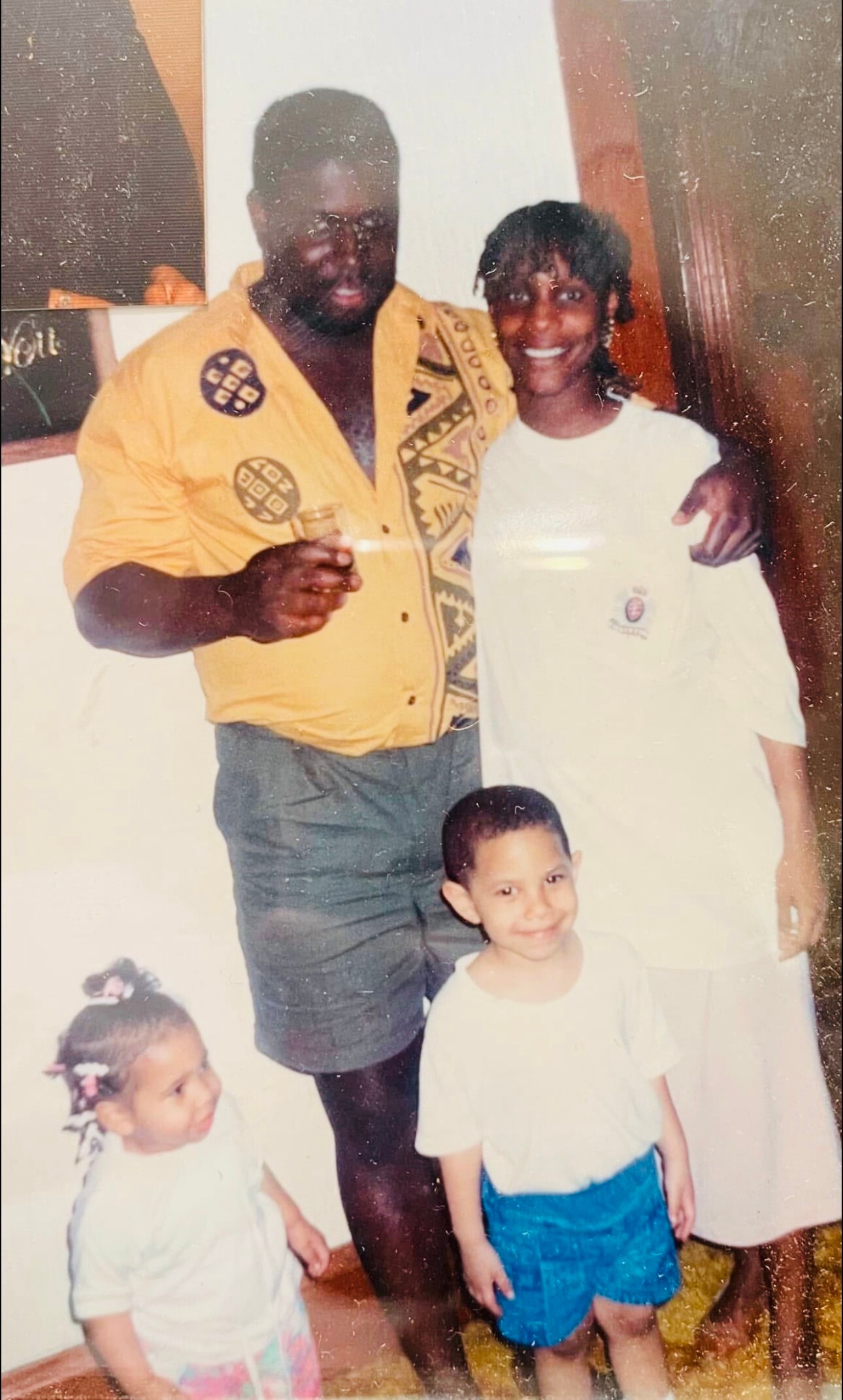 Father and mother with three young children posing together indoors; man in a yellow patterned shirt holding a drink, woman in white beside him.