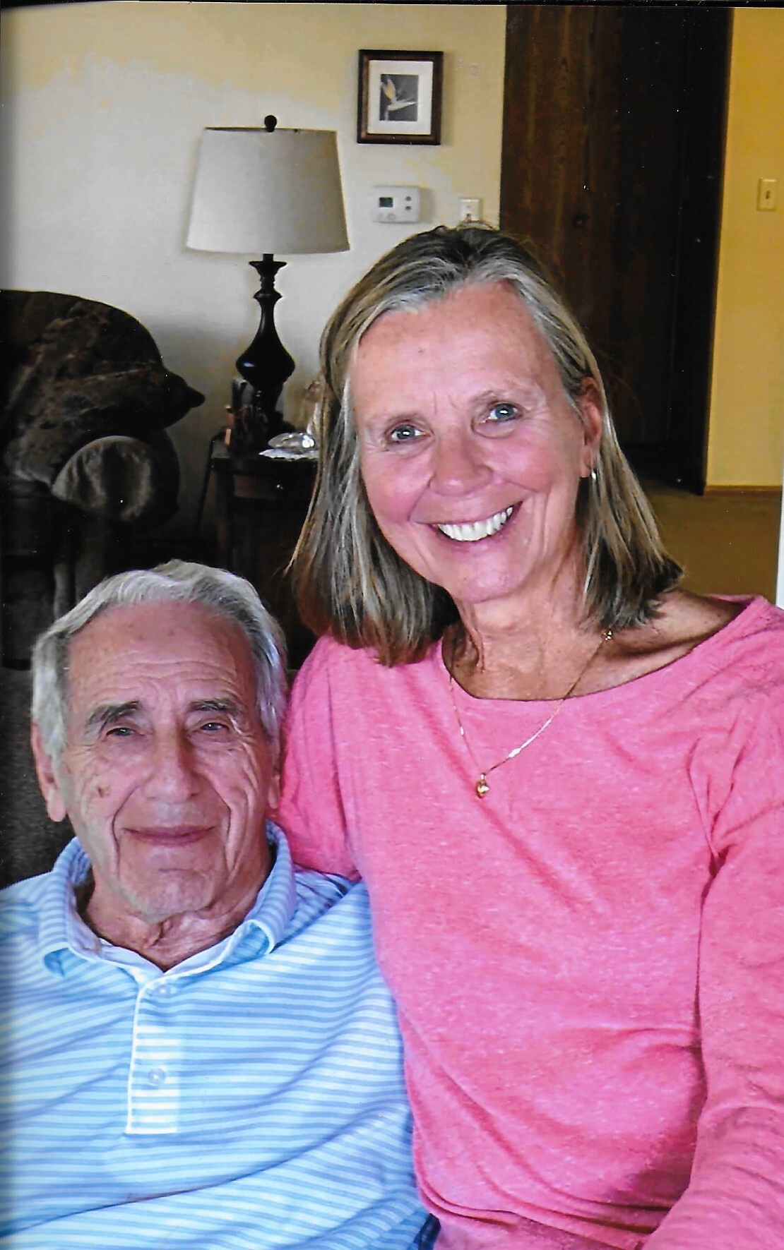 Smiling older couple sitting together in a living room, woman in pink top and man in blue striped shirt posing for a photo.