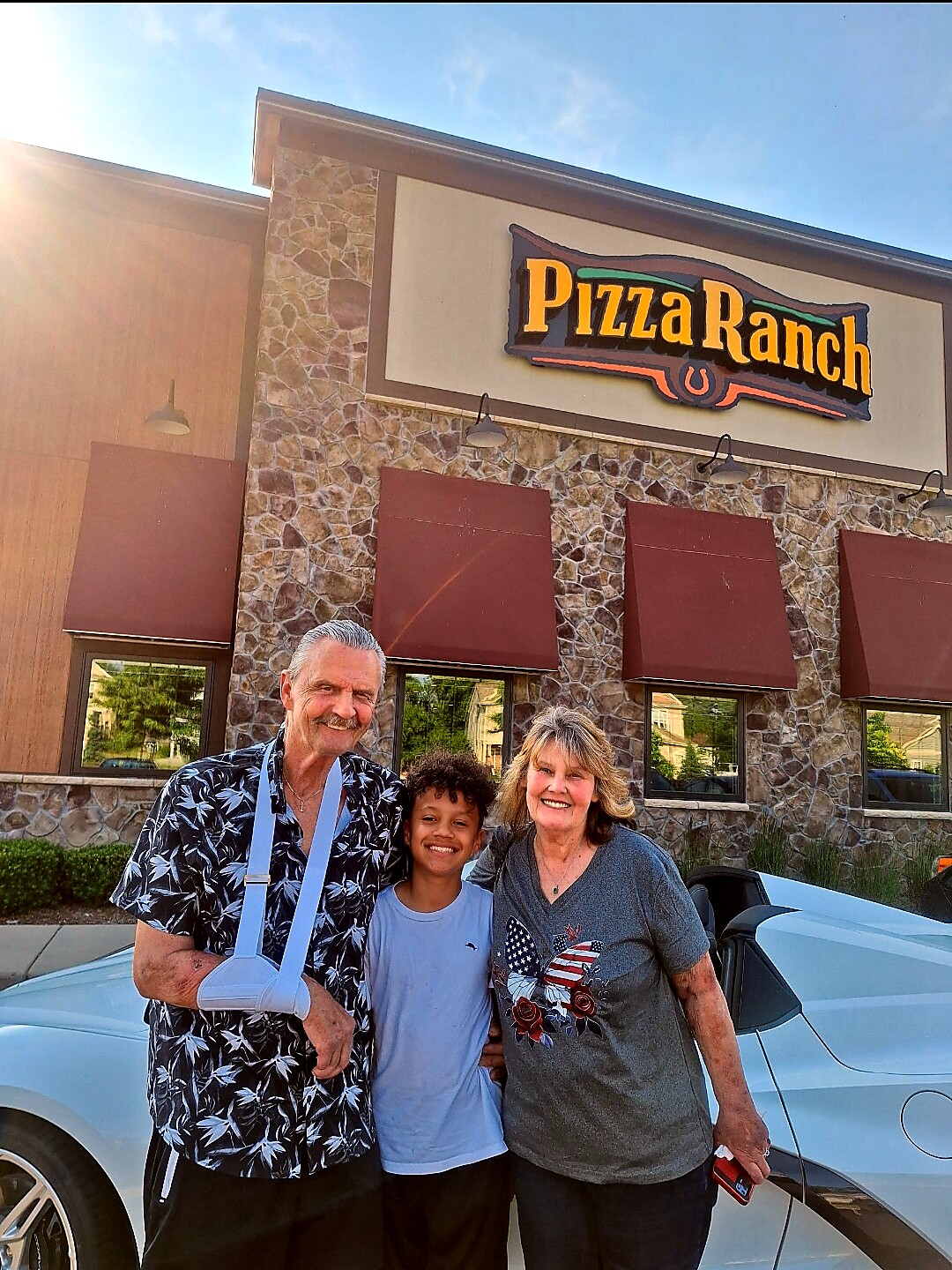 Three people stand together smiling in front of a Pizza Ranch restaurant, with a light blue car visible on the right and the stone façade behind them.