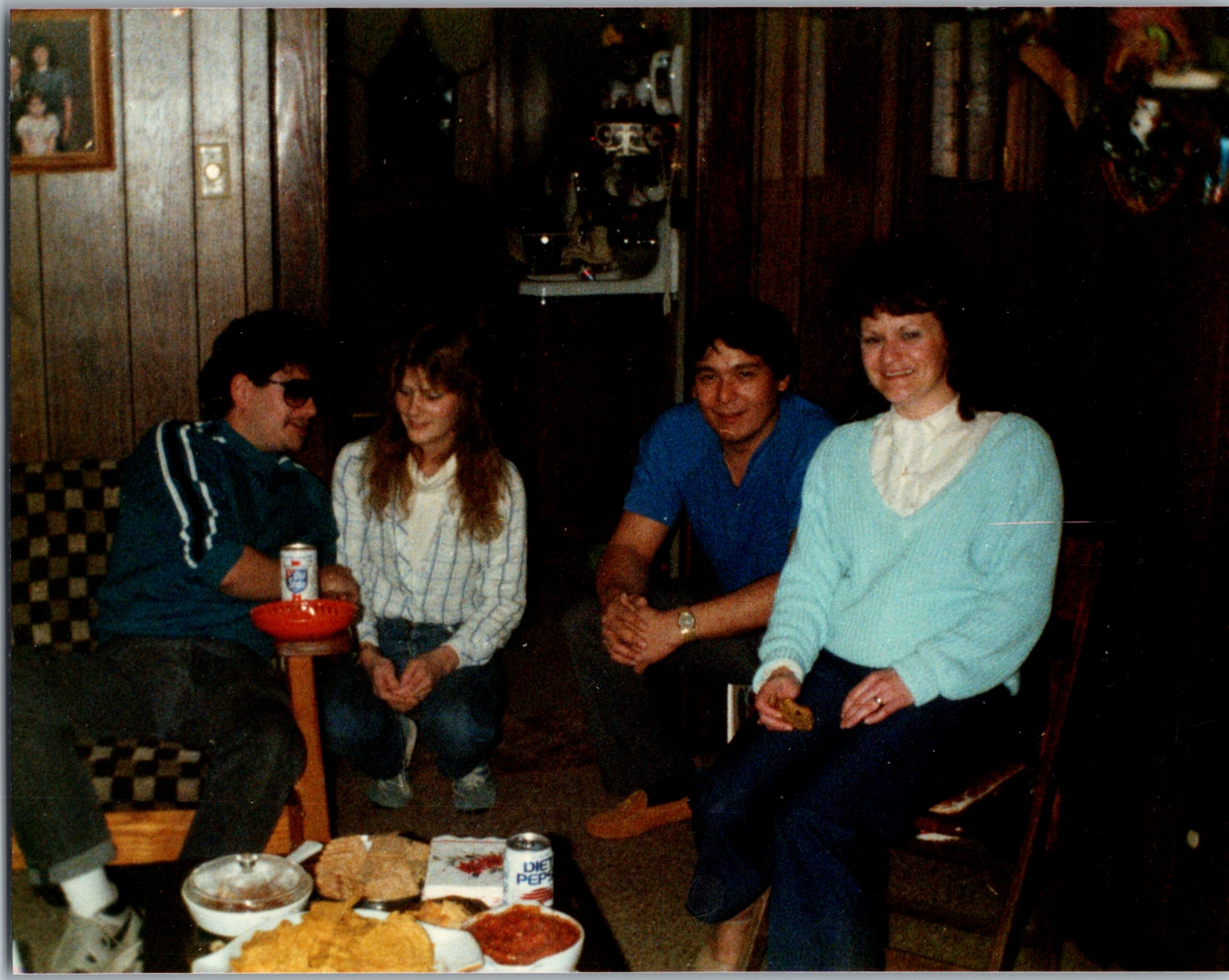 Four people sit around a table with bowls of chips and a Diet Pepsi can, chatting in a dim, retro dining room.