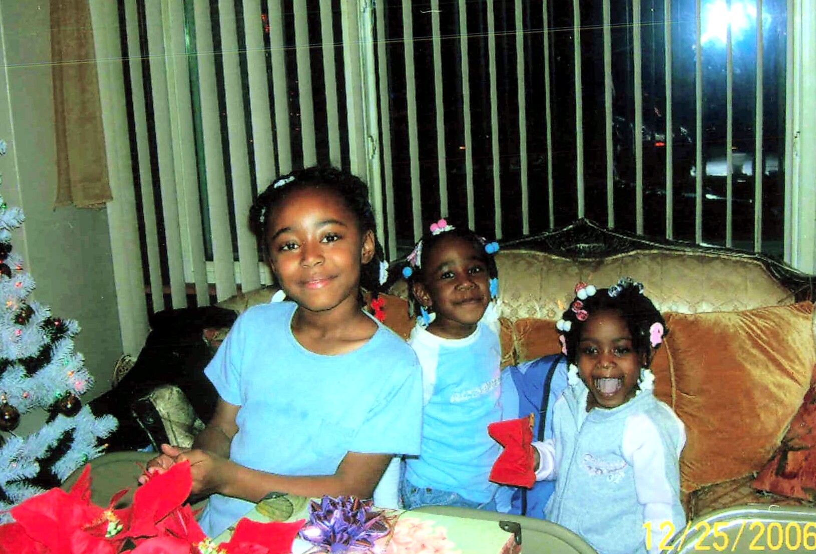 Three girls in blue shirts sit on a sofa, smiling, with a Christmas tree and wrapped gifts nearby.