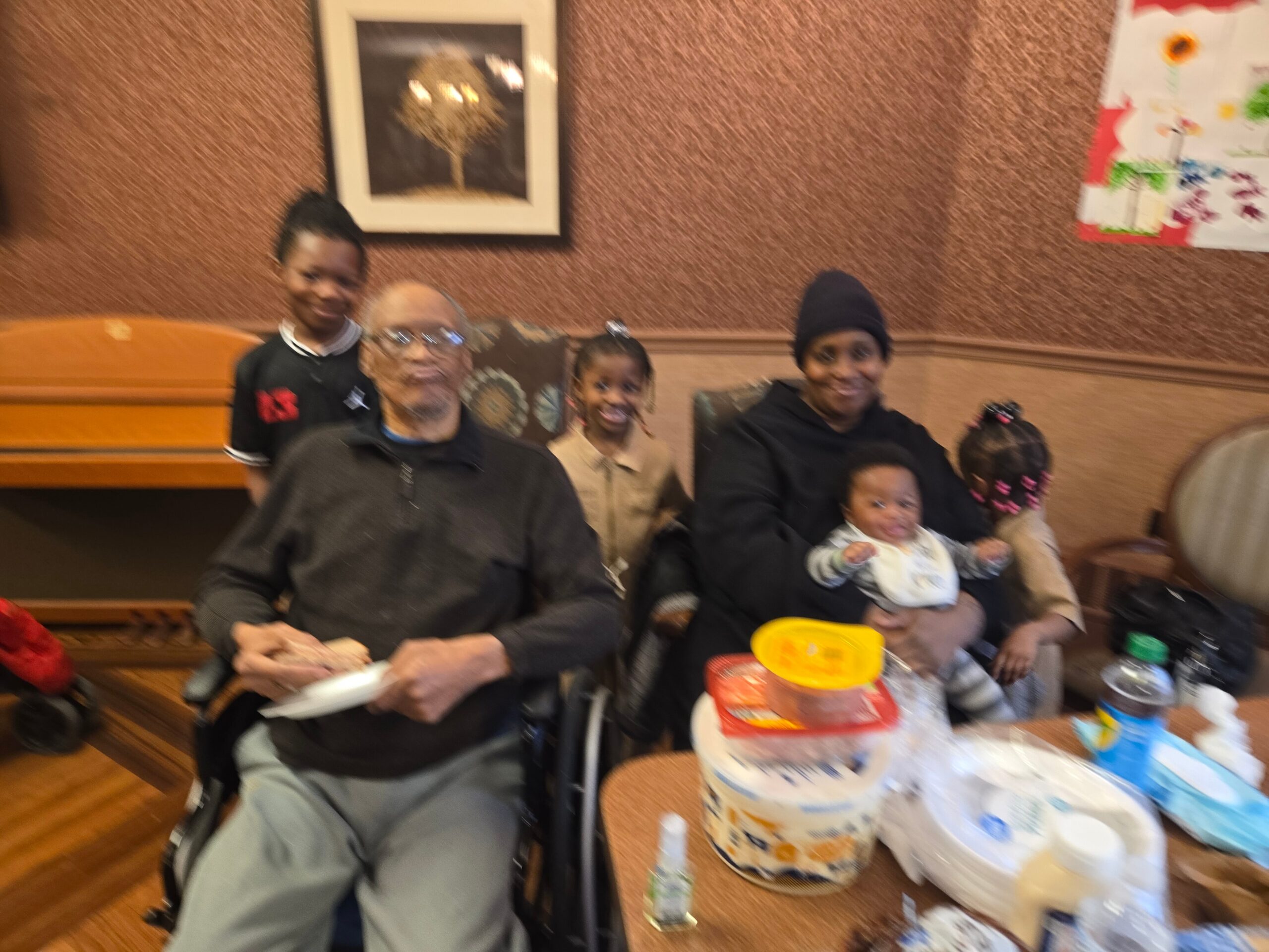 Group of family and friends at a birthday party, smiling around a table with a layered cake and party items in a warm room.