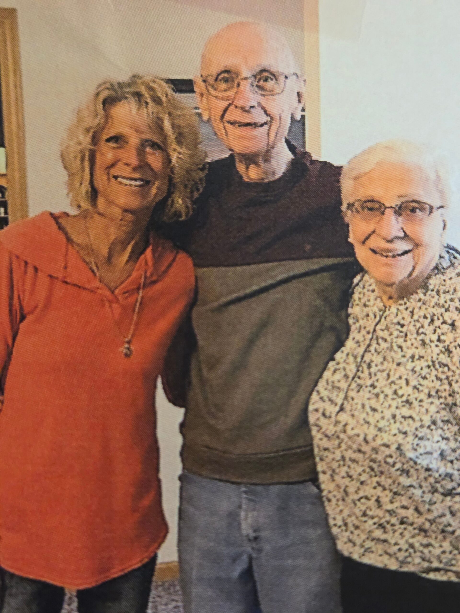 Three smiling adults posing together indoors for a photo, with a woman in orange on the left, a bald man with glasses in the middle, and a woman in a patterned sweater on the right.