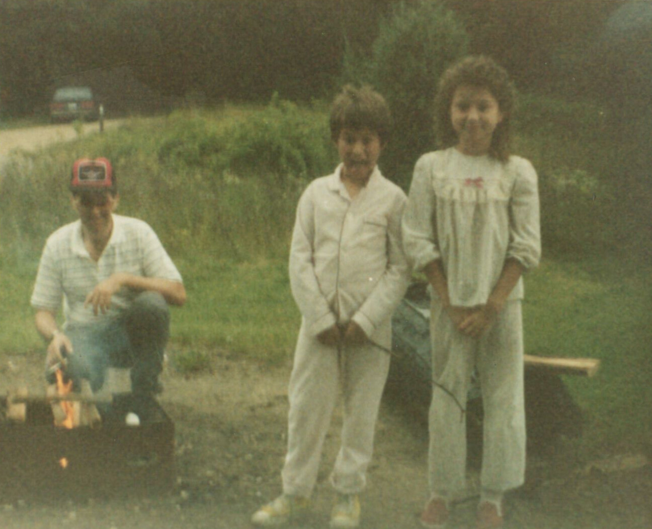 Three people pose outdoors in a grassy area: a man kneels to the left, while a boy in light clothing and a girl in a long dress stand side by side in the center.