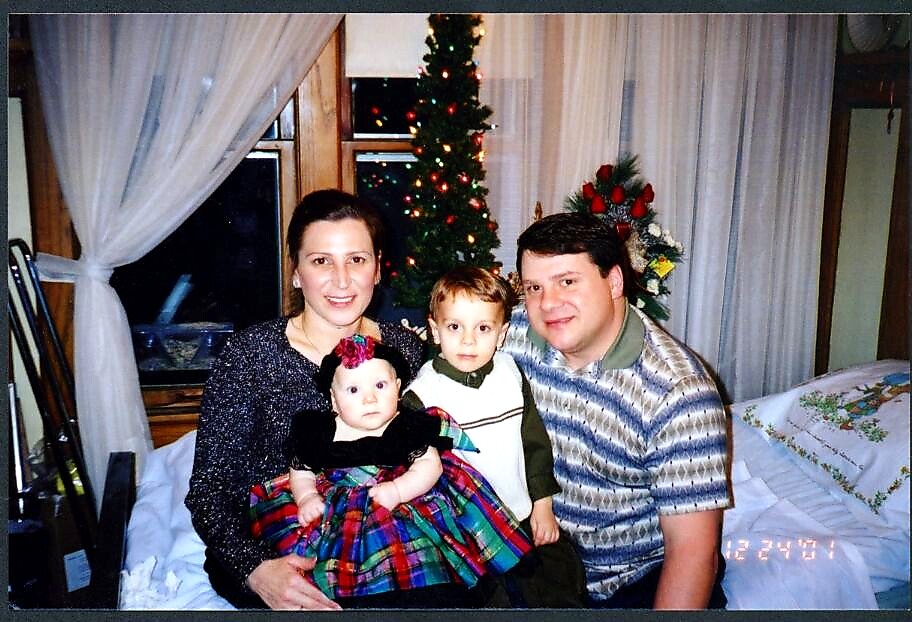 Family portrait of parents with two young children on a bed, Christmas tree and decorations in the background.