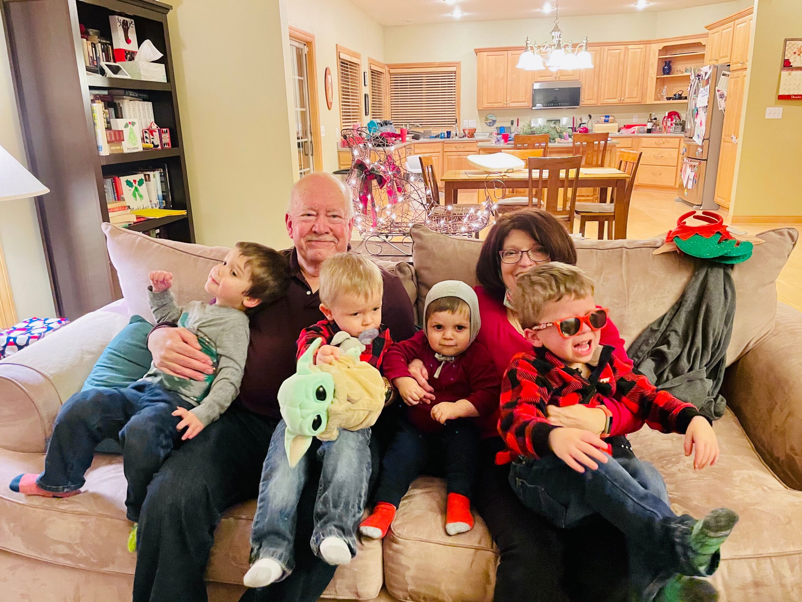 Family sitting on a beige sofa in a bright kitchen-living room, kids in festive clothes and a dragon hat visible on the armrest.
