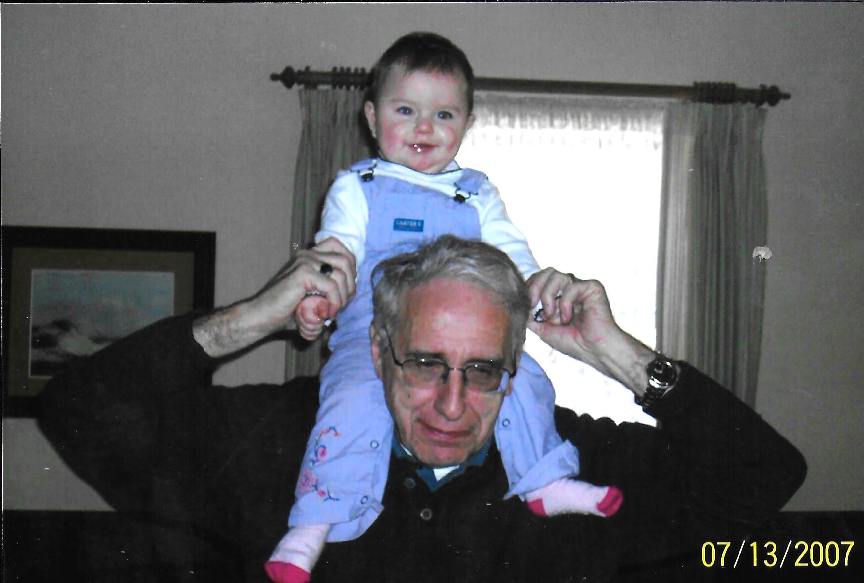 Grandfather wearing glasses lifts a smiling baby onto his shoulders indoors in a living room.