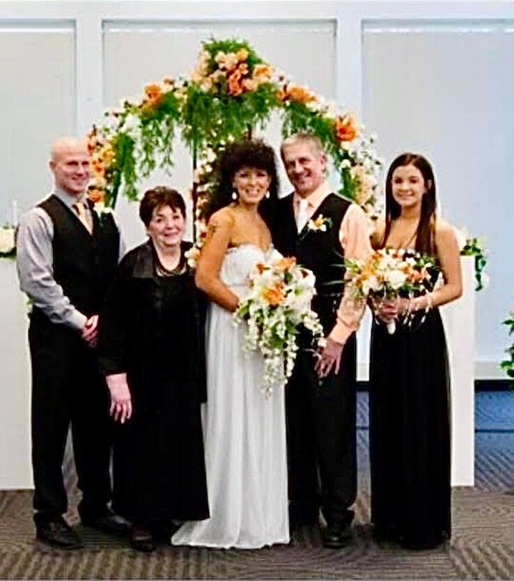 Wedding party posing indoors under a flower arch: bride in white gown holding a cascading bouquet, groom in vest and tie, a man in black vest, and two women in black dresses with bouquets.