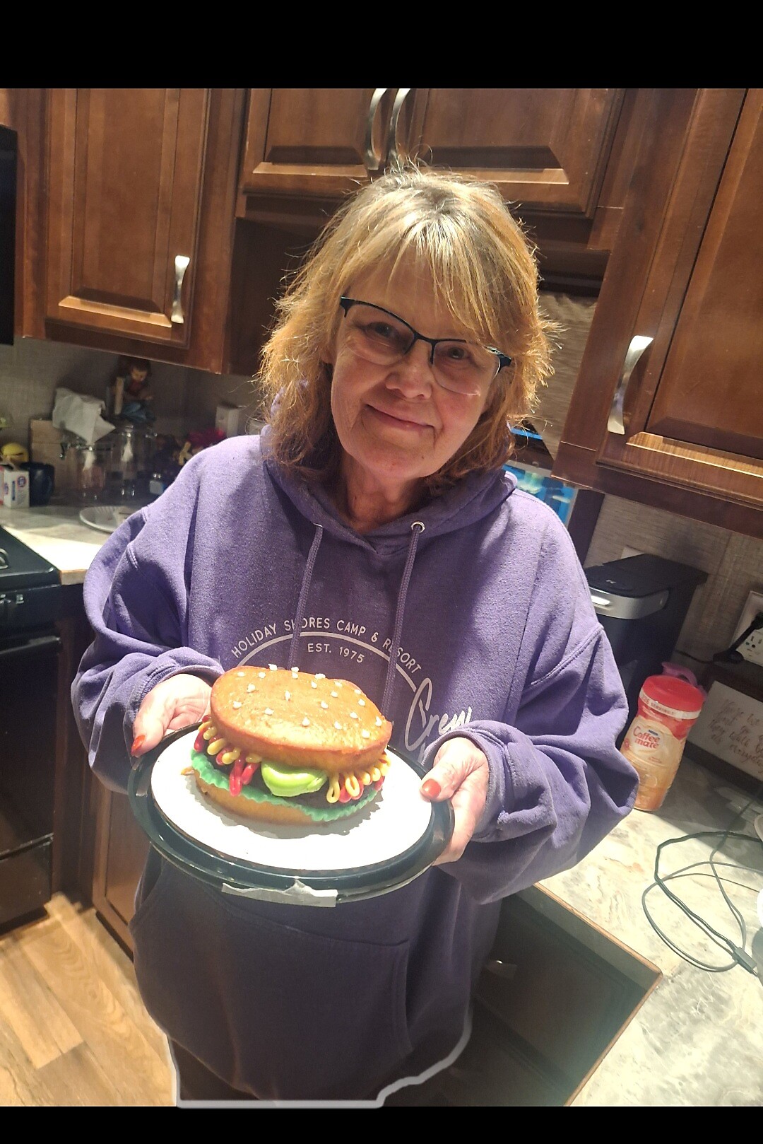 Smiling older woman in a purple hoodie presenting a hamburger-shaped cake on a plate in a kitchen with wooden cabinets behind her.