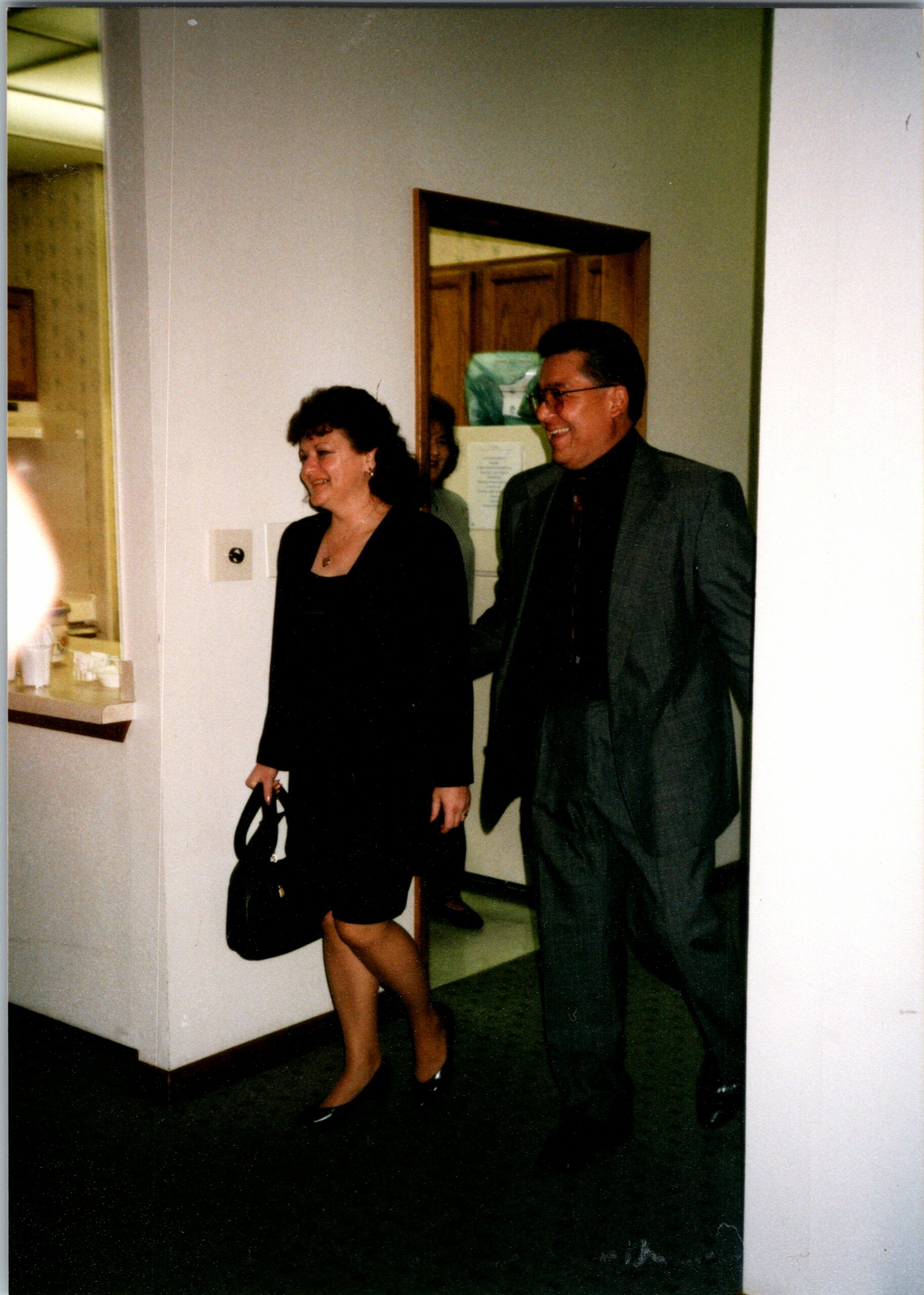 Smiling man in a gray suit and a woman in a black dress walk down a hallway indoors with a handbag.