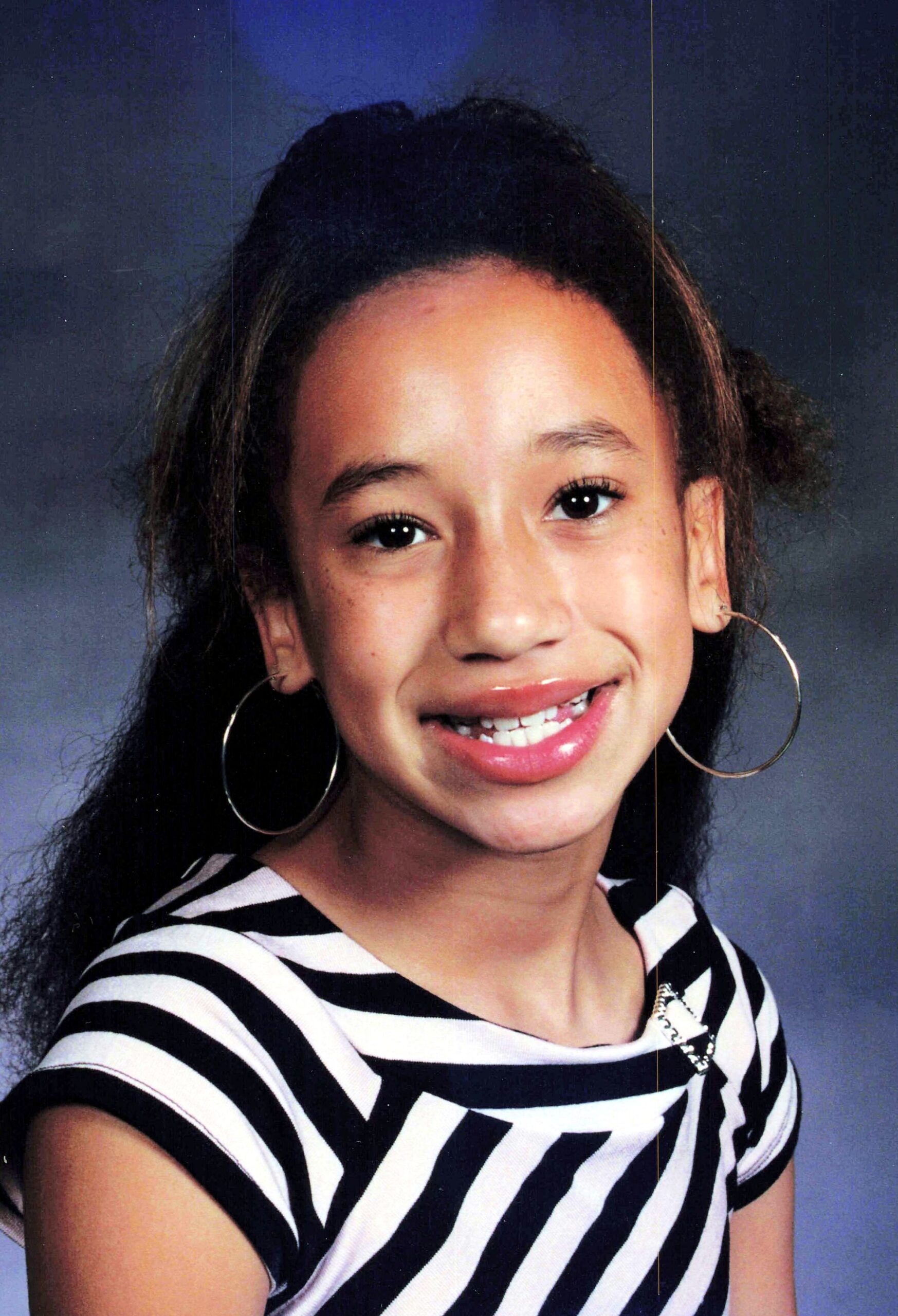 Smiling young girl with long dark hair, wearing a black-and-white striped shirt and large hoop earrings, portrait against a blue backdrop.
