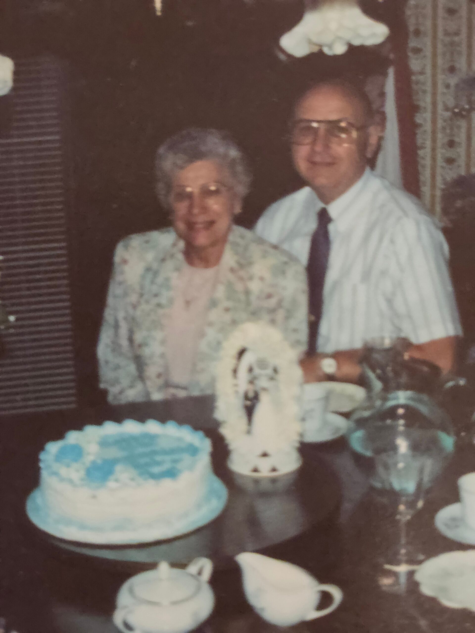 An elderly couple smiles at a celebration, with a blue-and-white frosted cake and teacups on the table in front of them.