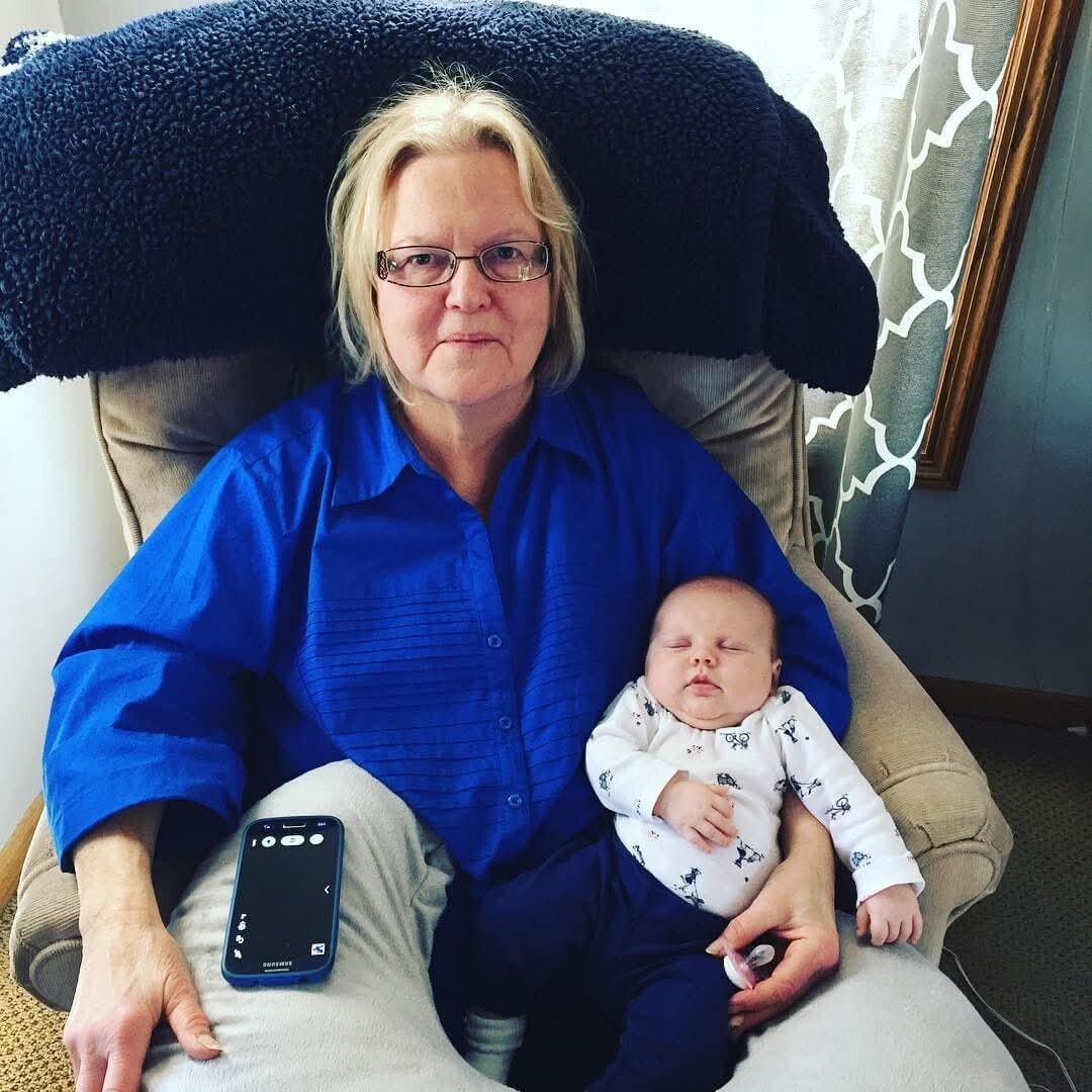 Older woman with short blonde hair and glasses sits in a recliner wearing a blue shirt, holding a sleeping baby in a white onesie on her lap; a smartphone rests on her leg to the side.
