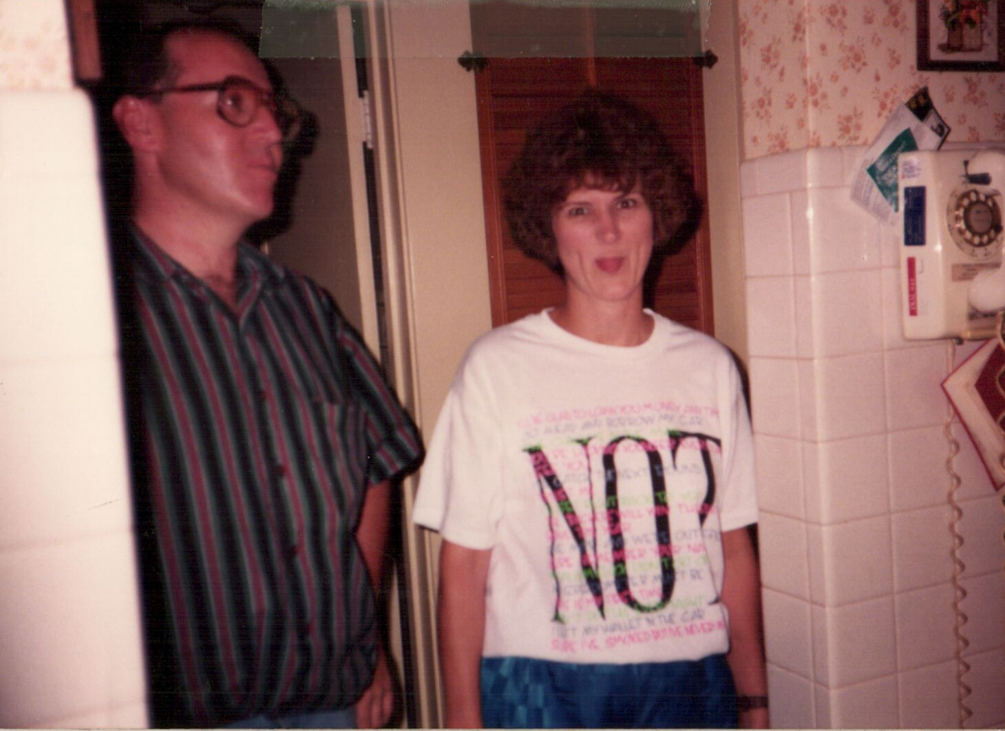 Two adults stand in a retro kitchen; man in a striped shirt on the left, woman in a white graphic tee on the right, both looking at the camera and smiling.