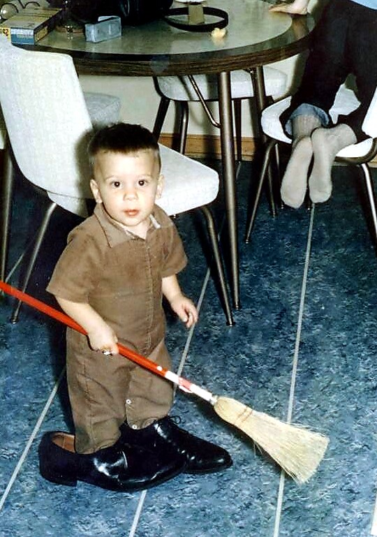 Toddler in a brown outfit standing on a blue floor, sweeping with a small broom and wearing oversized black shoes.