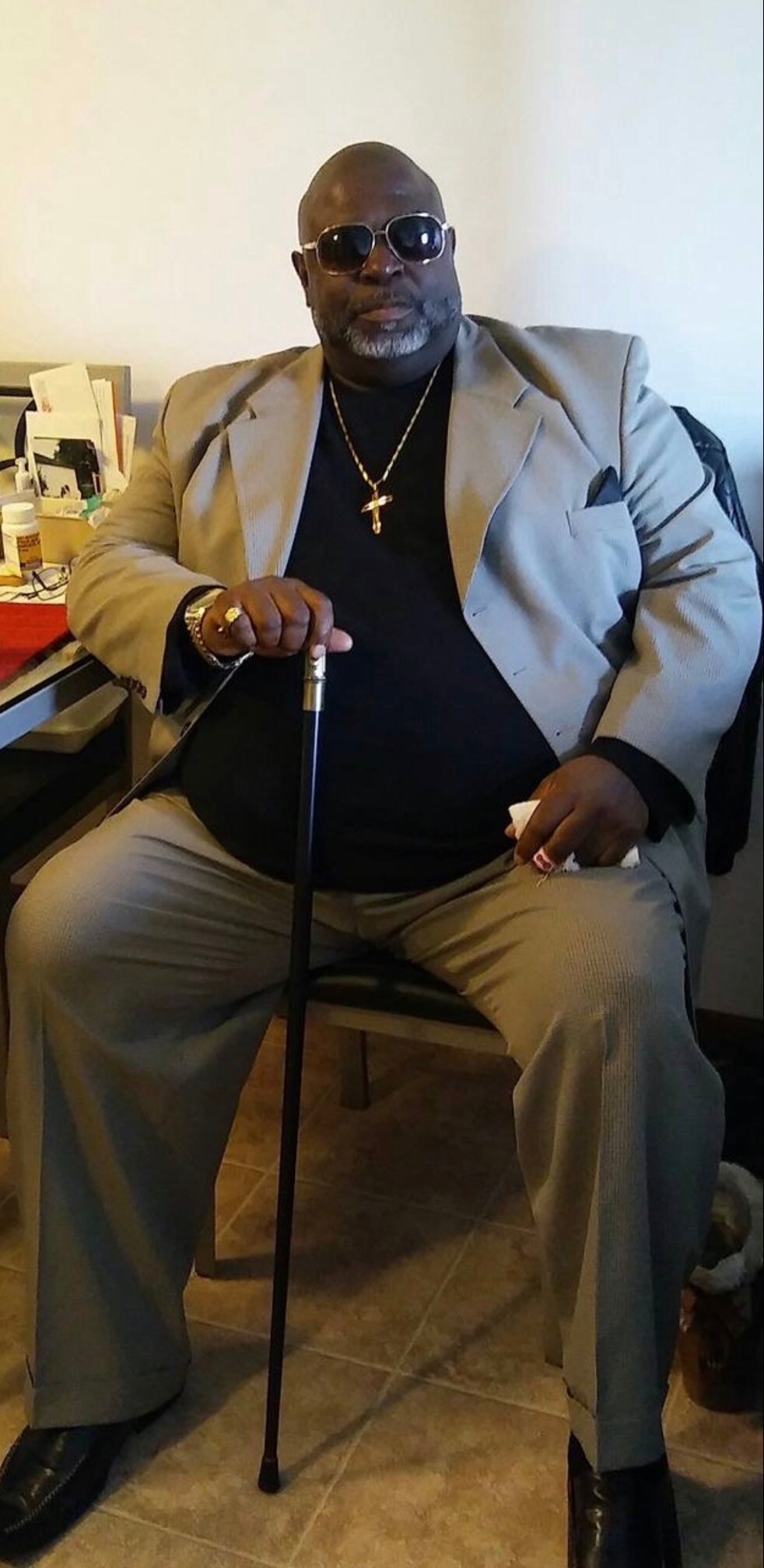Man in a beige suit and black shirt sits with a cane, wearing sunglasses and a gold cross necklace.
