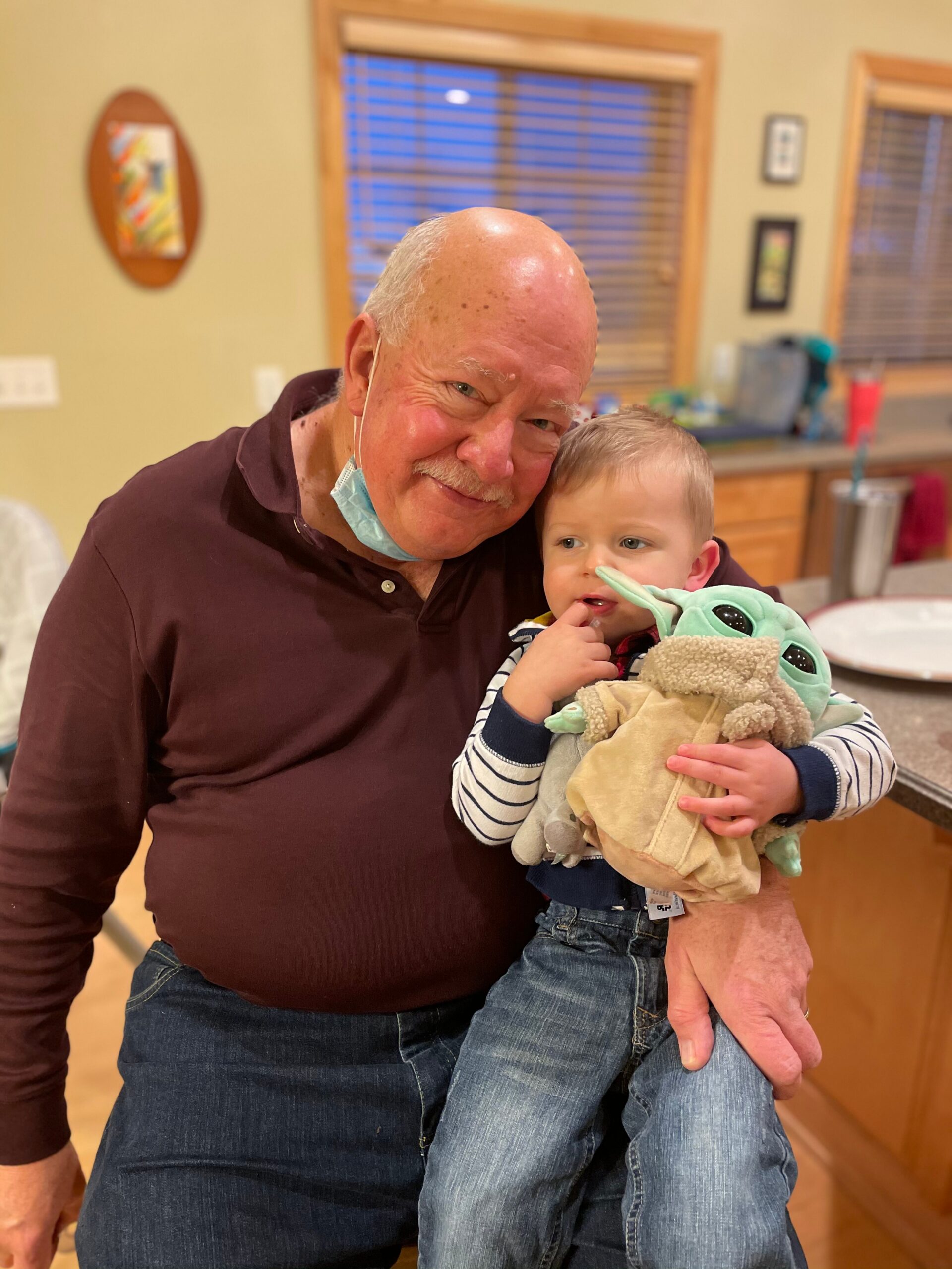 Grinning older man holding a baby in a frog-themed hoodie in a kitchen setting.