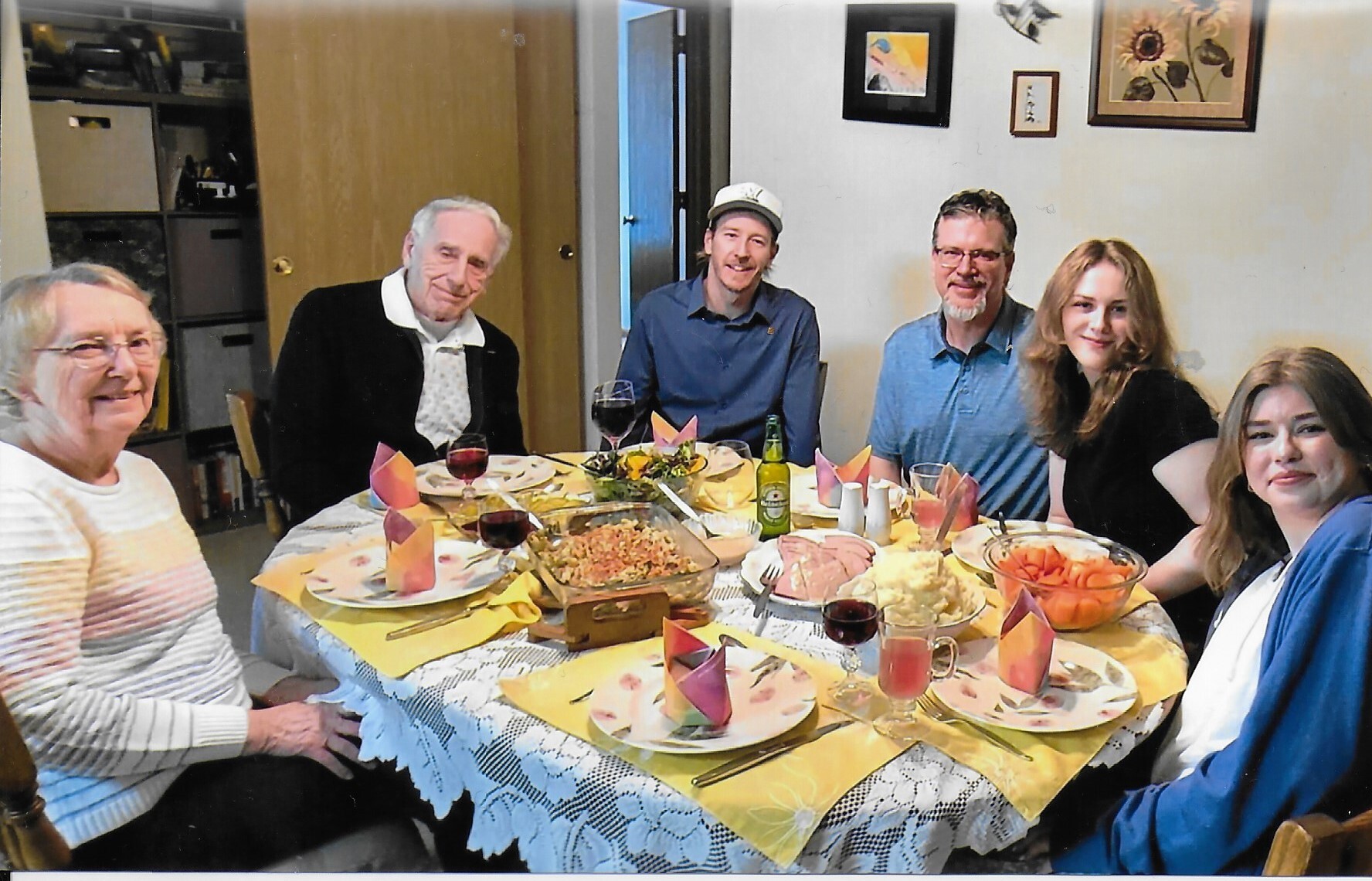 Six family members sit around a table enjoying a meal, with dishes, wine, and a bottle visible on the table.