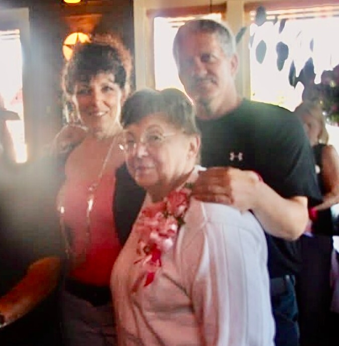 Three adults posing indoors; older woman center wearing a pink lei, flanked by a woman in coral top on the left and a man in a black shirt on the right.