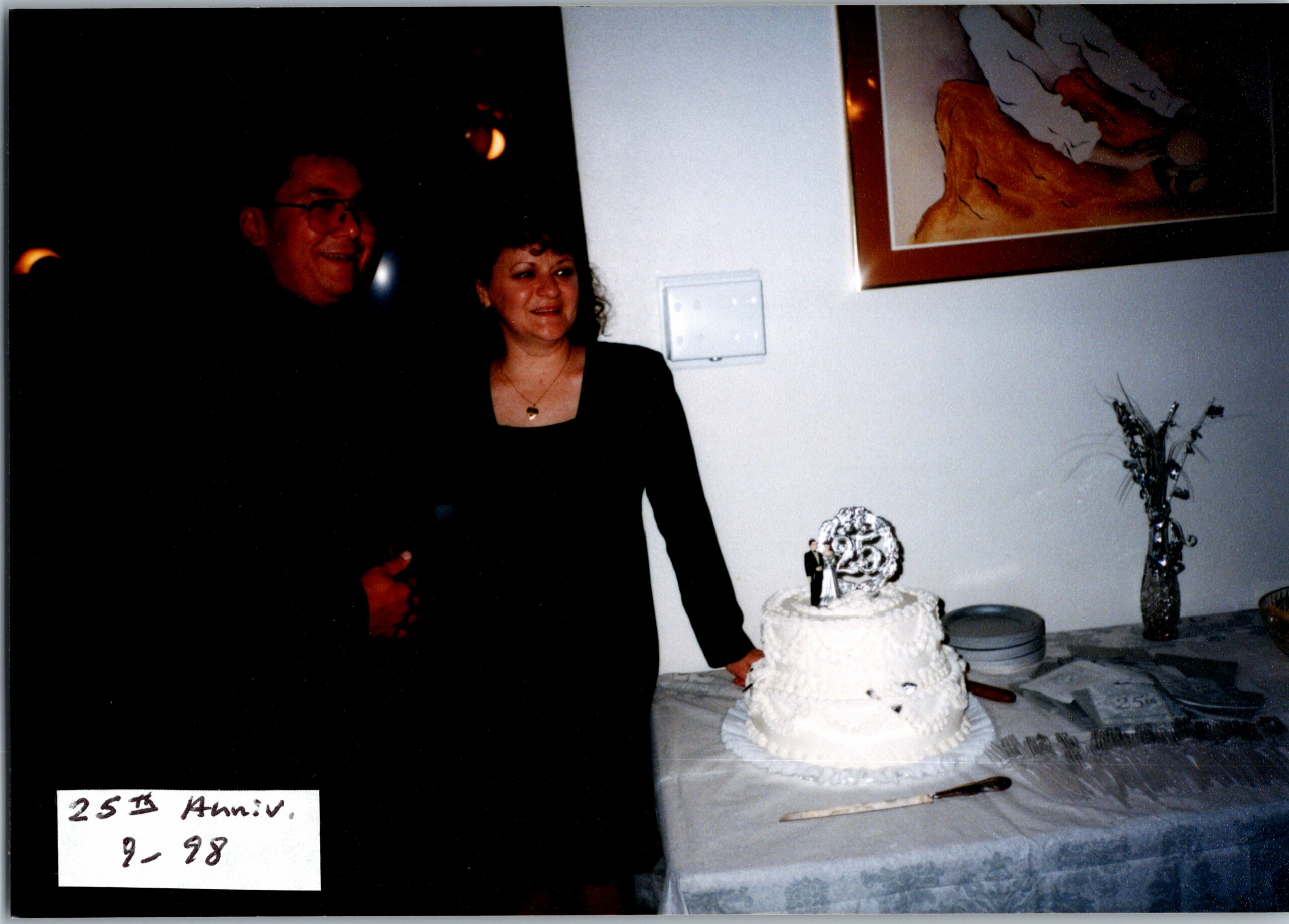Woman in black stands beside a two-tier white cake at a 25th anniversary party, smiling at the table.