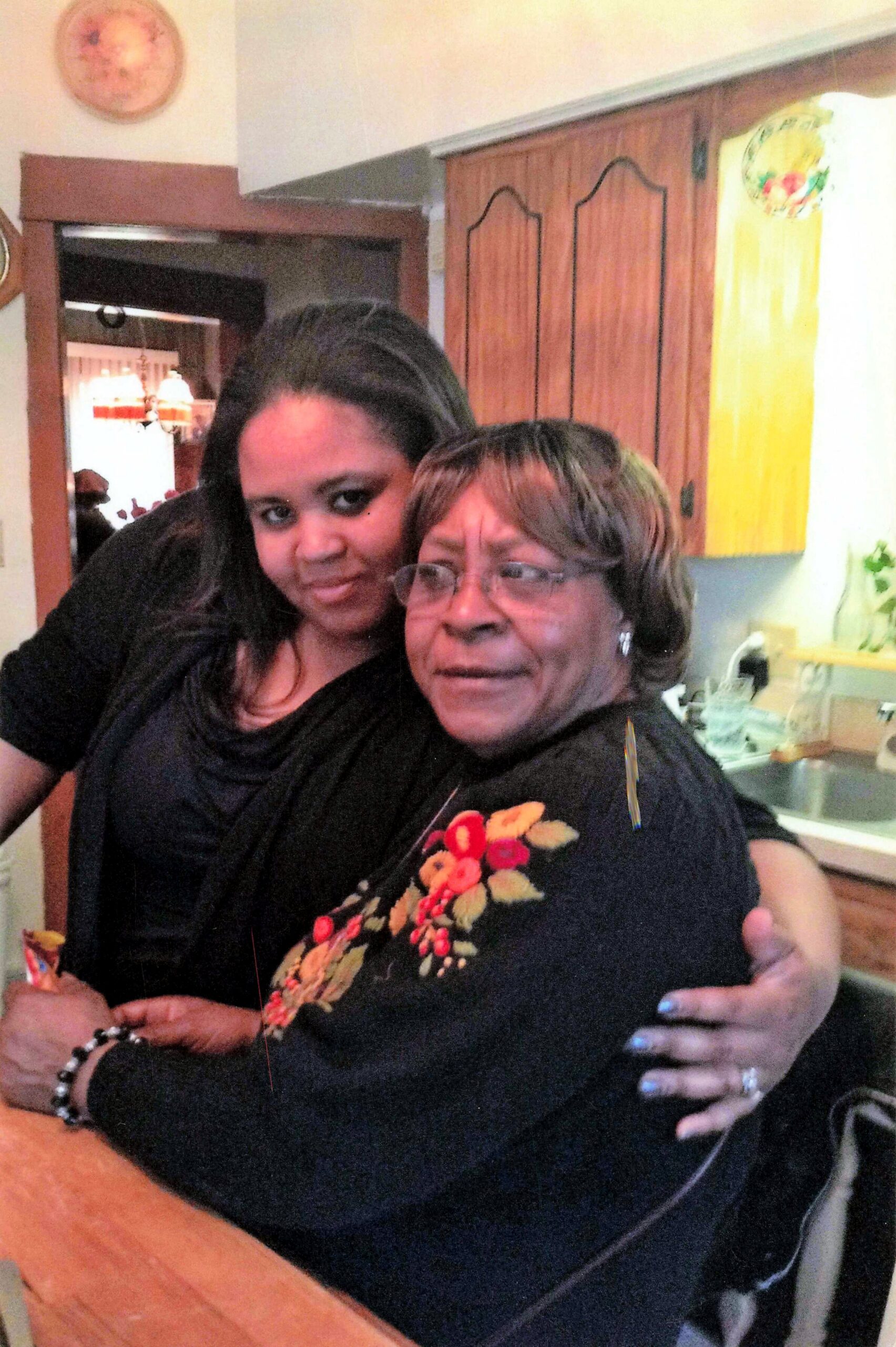 Two women hug for a photo in a kitchen, smiling at the camera with one wearing glasses and a black embroidered top.