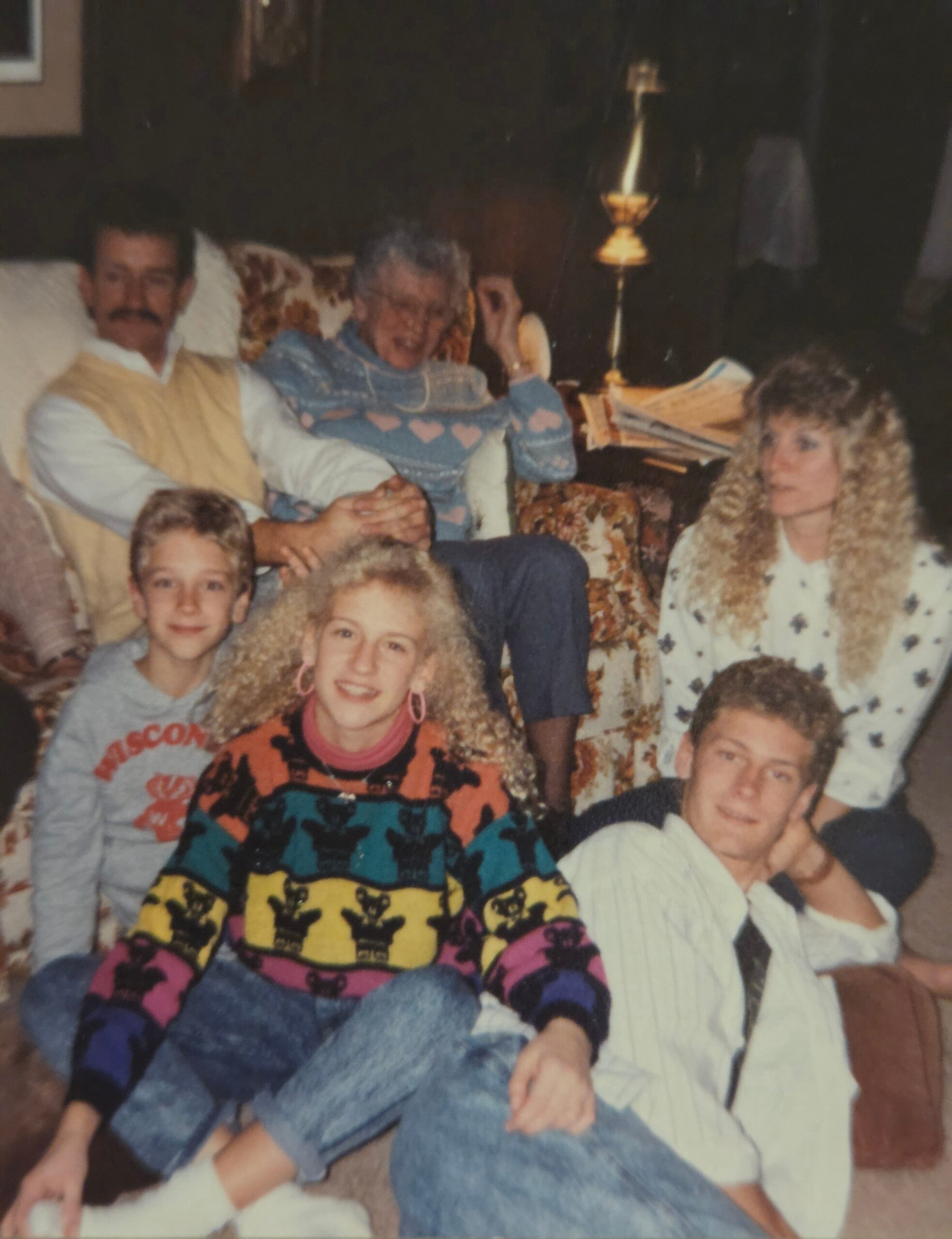 A family photo in a living room: children sitting on the floor and adults on a floral couch, all smiling in colorful, retro sweaters.