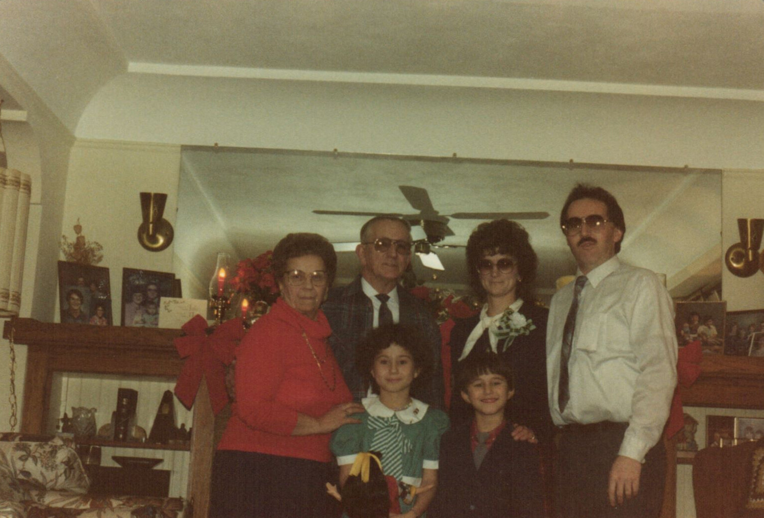 Five-family members pose for a holiday photo in a decorated living room with a fireplace mantel and red ribbons/flowers in the background.