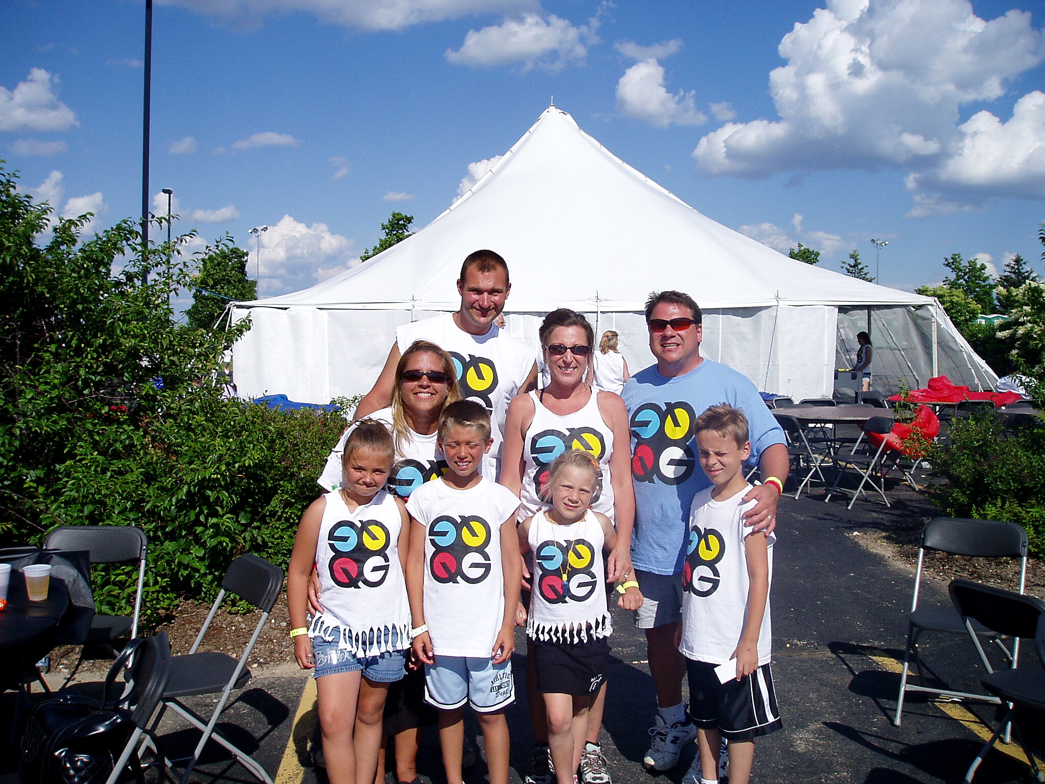 Group of smiling people in white shirts with a colorful circular logo posing outdoors in front of a large white event tent.