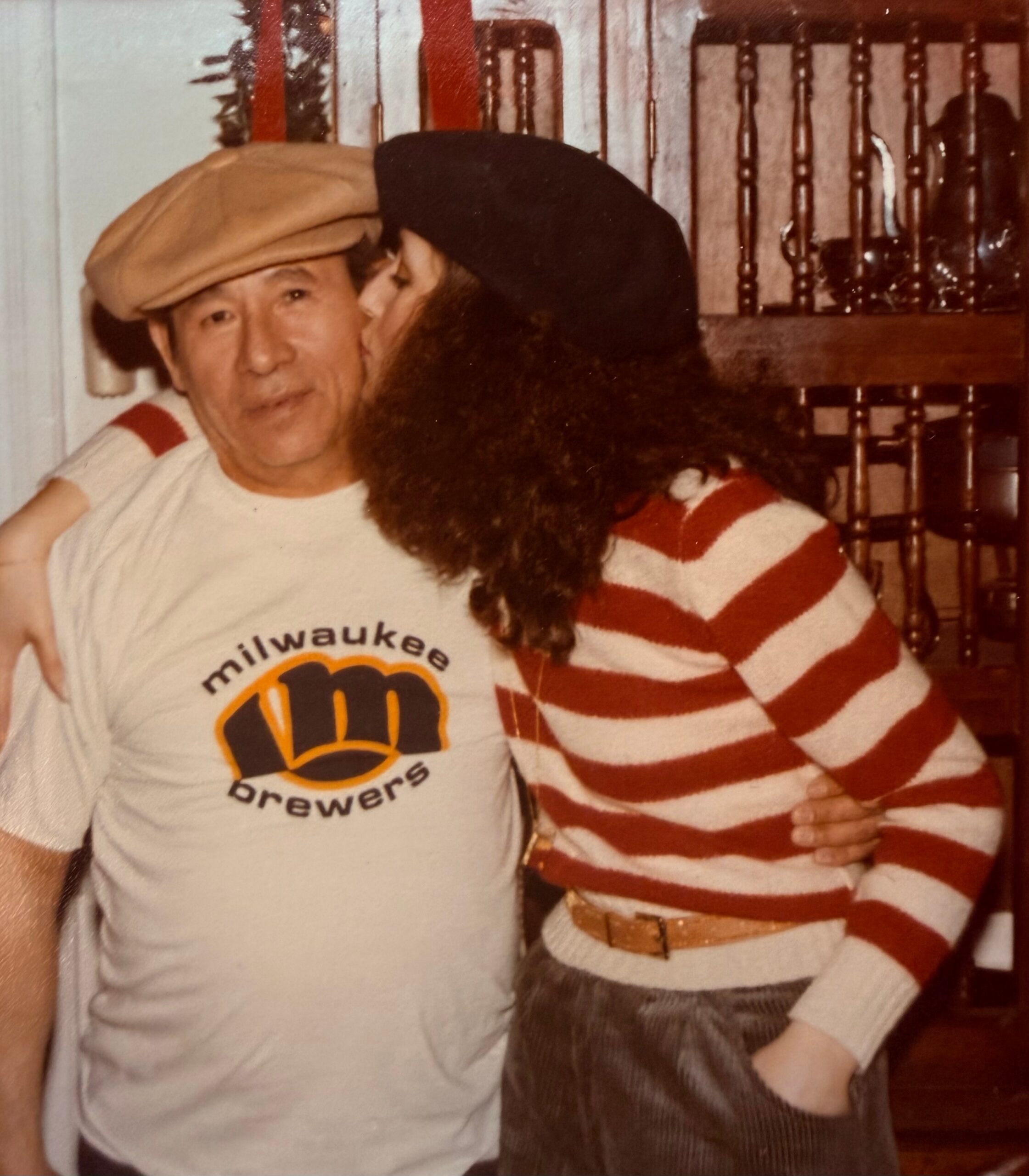 Man in a Milwaukee Brewers shirt and tan cap smiles as a friend hugs him indoors.