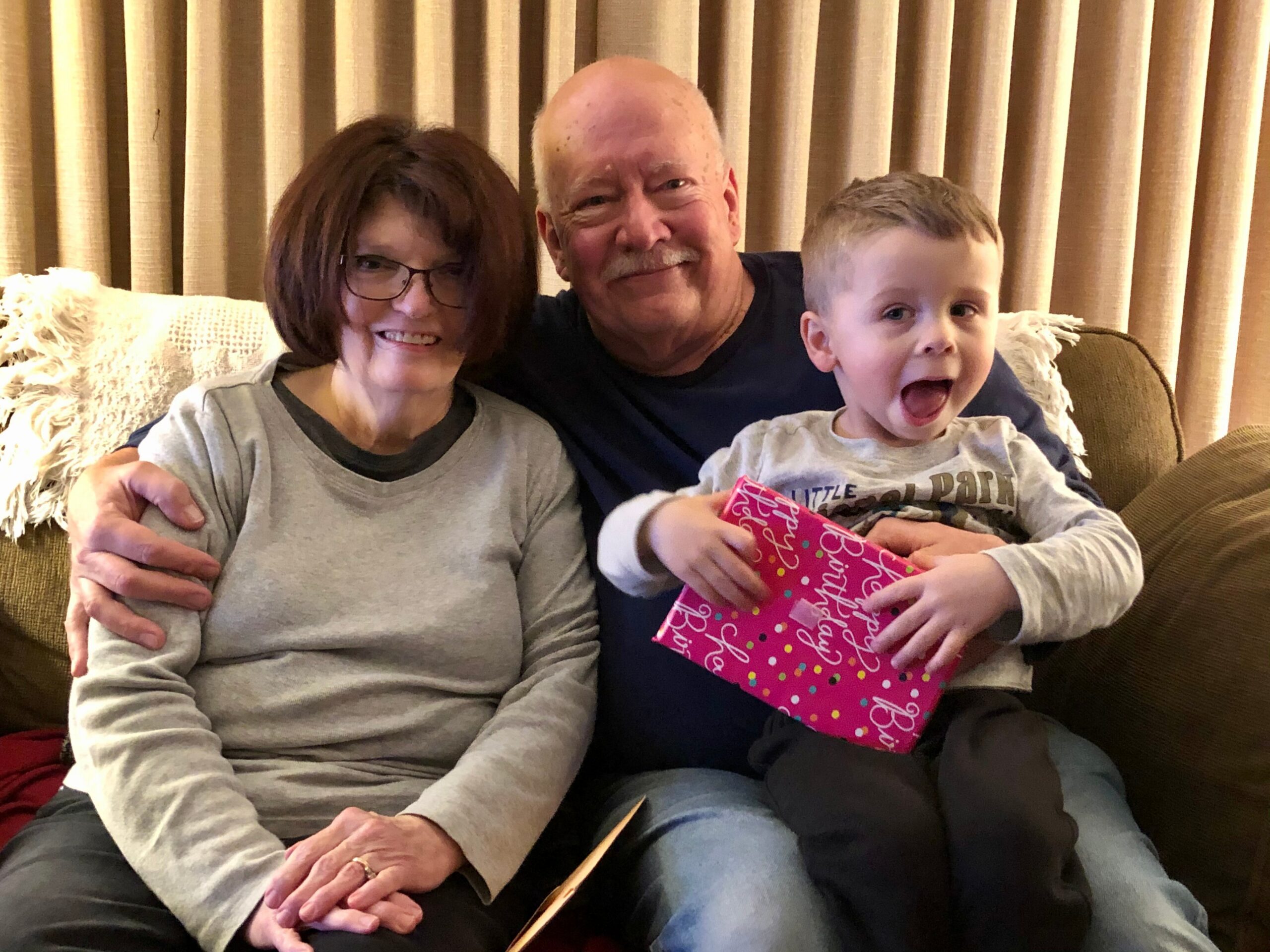 Three generations posing on a sofa: grandmother with glasses, grandfather in the middle, and a smiling toddler holding a pink birthday gift.