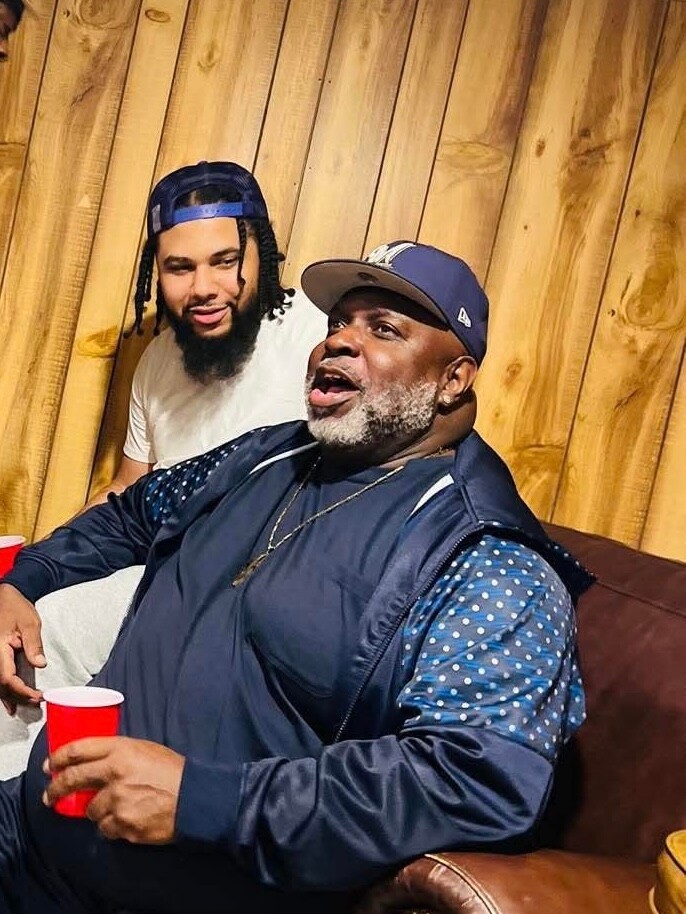 Two men sit on a brown couch in a wood-paneled room, laughing together; one holds a red cup.