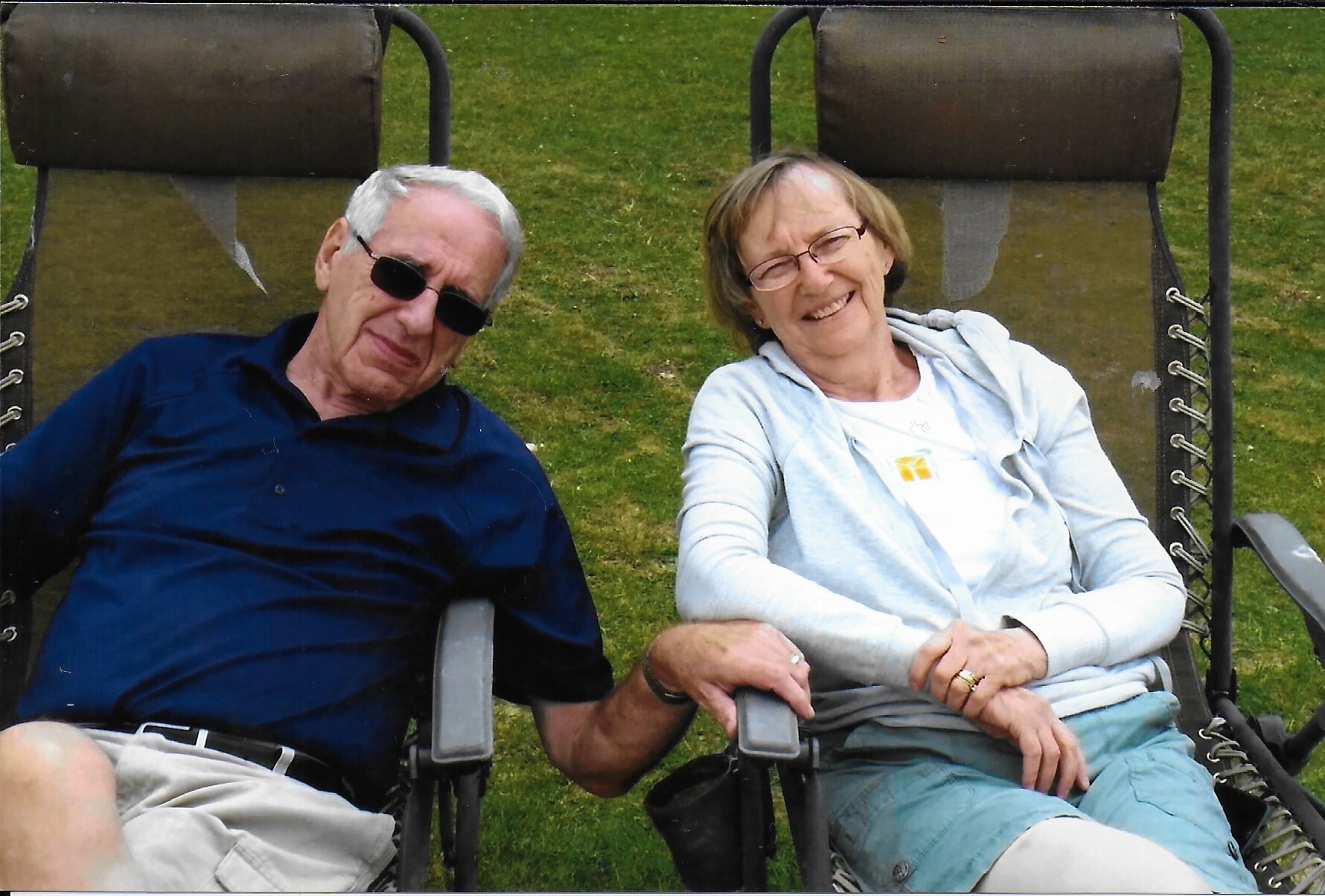 Older couple relaxing in outdoor lounge chairs on a grassy lawn, smiling at the camera.