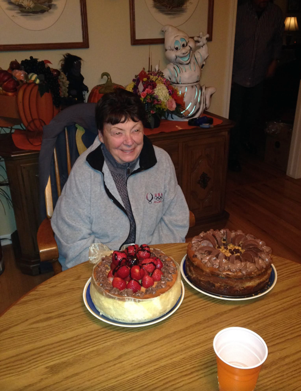 Smiling woman in a gray jacket sits at a wooden table with two cakes—one topped with strawberries and the other chocolate—in a cozy, decorated room with pumpkins in the background.