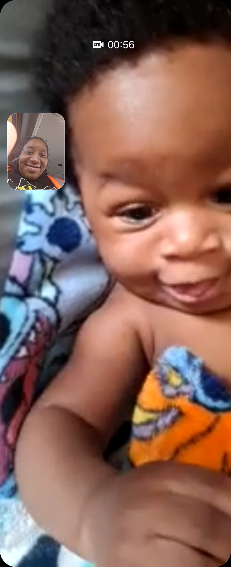 Close-up of smiling baby wrapped in a colorful towel, seen in a video call with an inset of a woman wearing an orange safety vest.