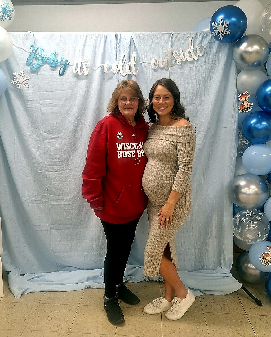 Pregnant woman in a beige knit dress posing with a friend in a red Wisconsin hoodie in front of a light-blue winter backdrop with balloons and a 'Baby it's cold outside' banner.