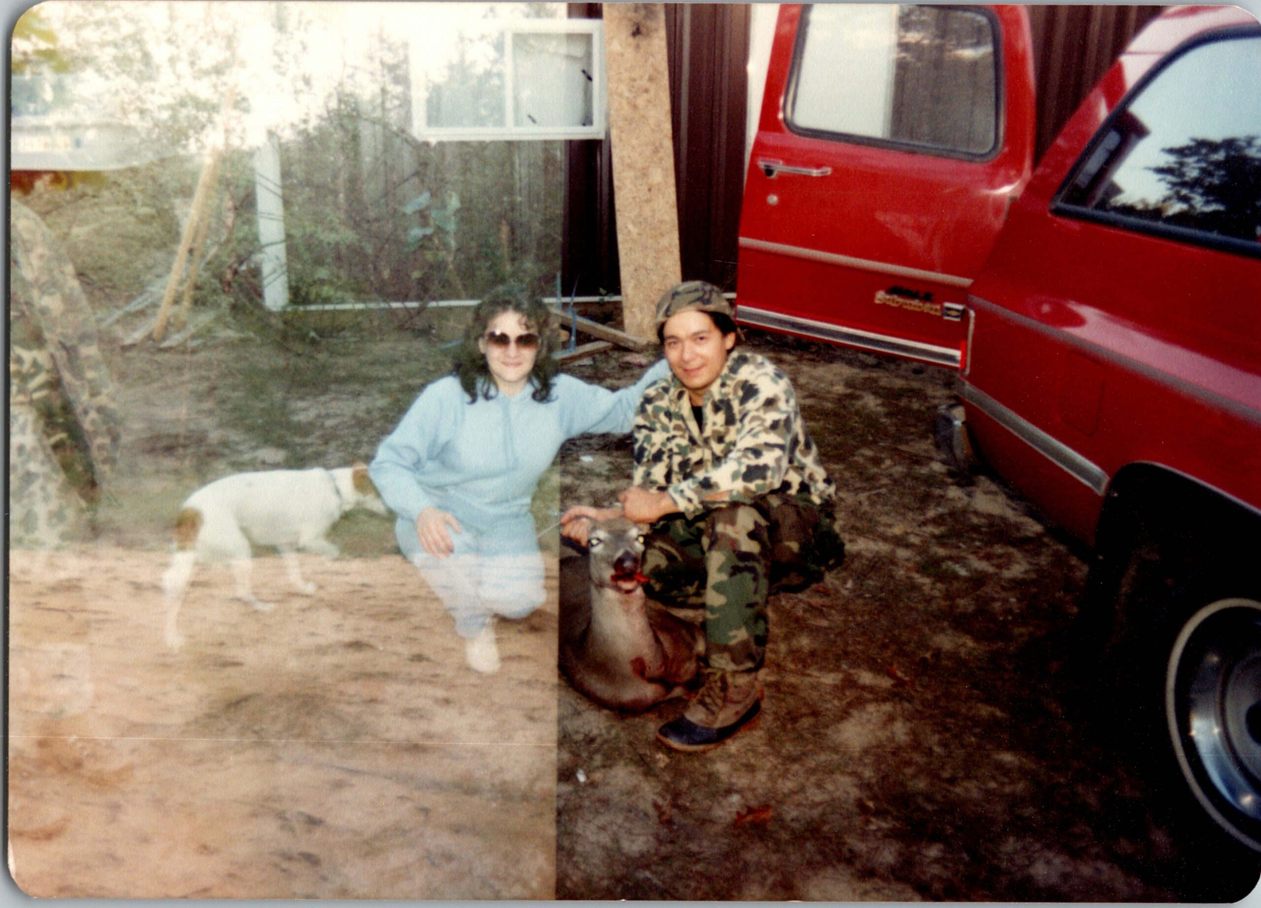 Two people sit on the ground near a red truck in an outdoor yard, with a white dog nearby; casual, taken during daylight by a casual photographer.