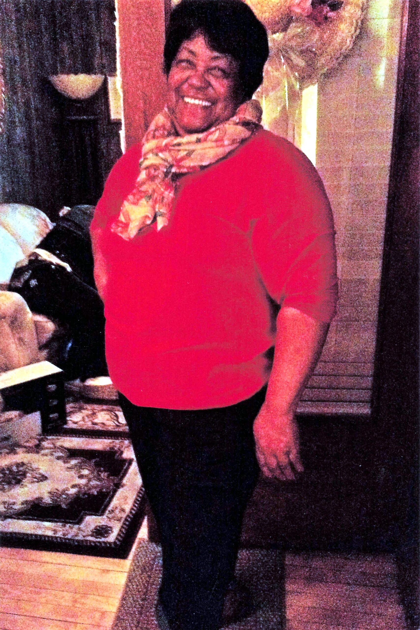 Smiling woman in a red blouse and floral scarf standing indoors near a glass cabinet or display case.