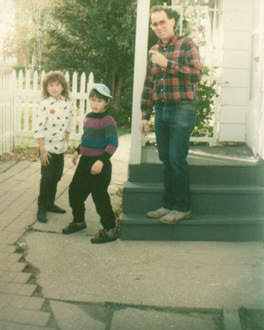 A man in a red plaid shirt stands on porch steps with two children in front yard, white picket fence behind them in daylight.