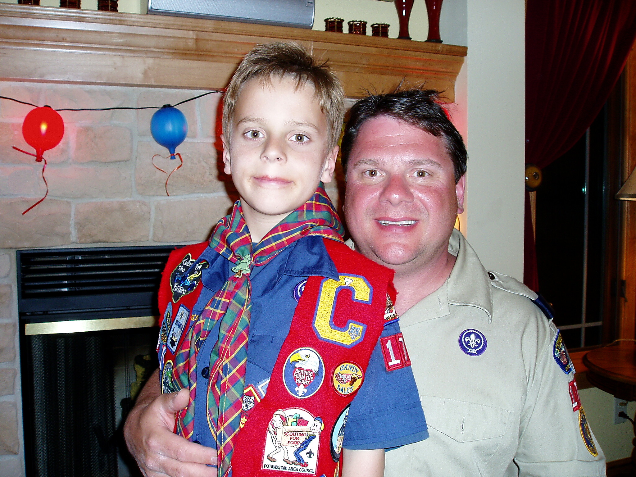 Father and son in scout uniforms posing for a photo indoors, with a red/blue balloon garland in the background and a stone fireplace behind them.