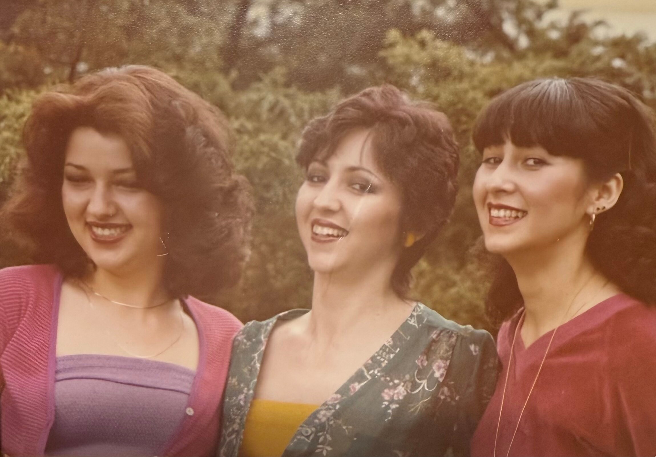Three women smiling and posing together outdoors, with trees in the background, looking at the camera.