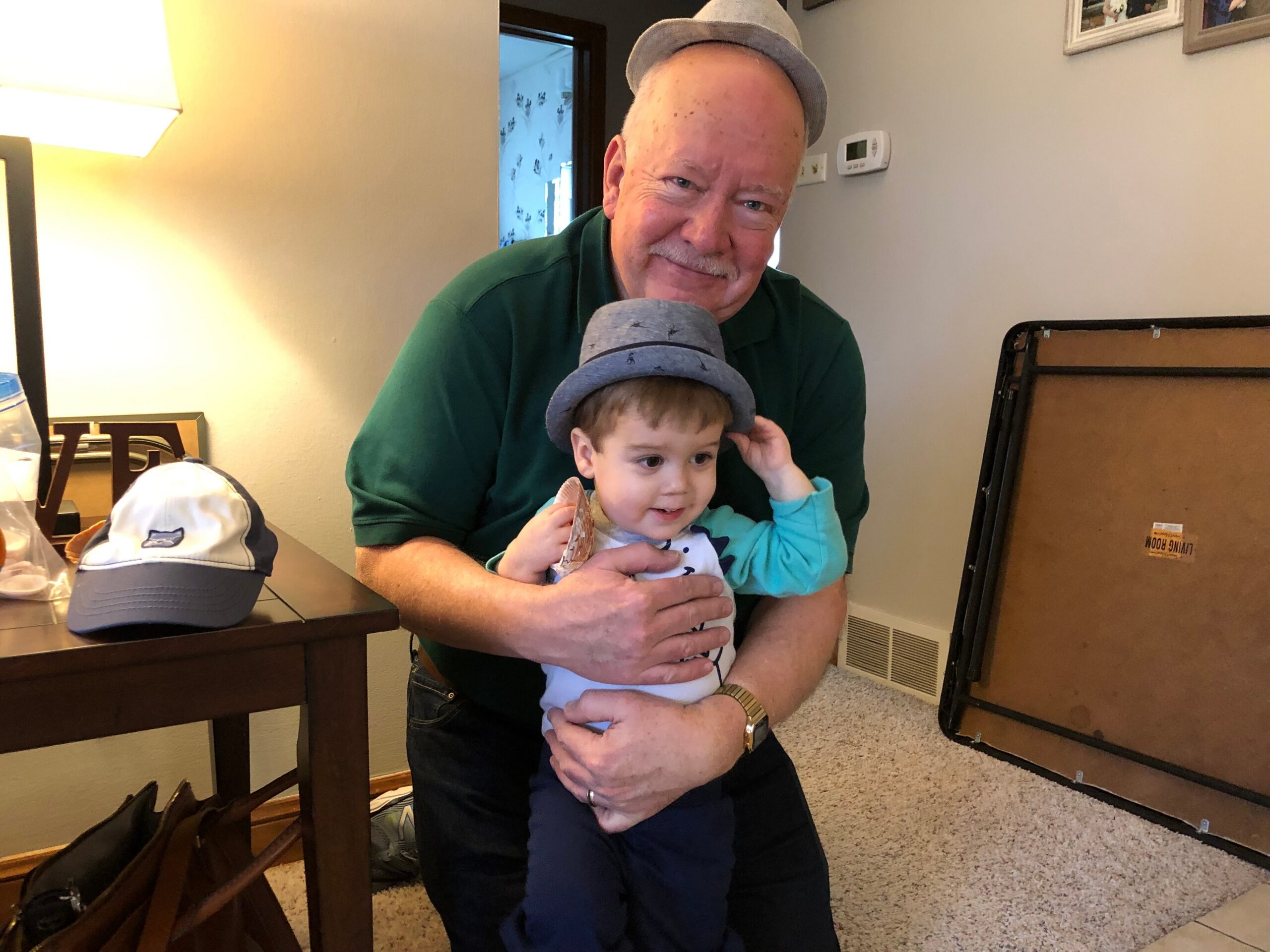 Smiling man in a green shirt holding a small child wearing a gray hat indoors, near a table and a suitcase-like object in the background.
