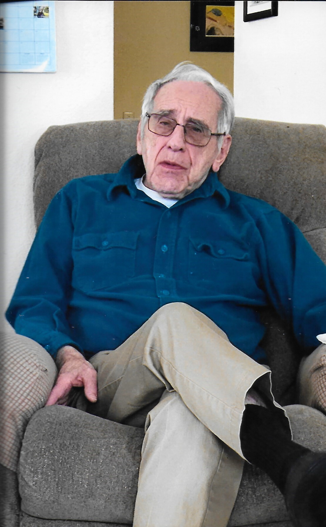 Older man with gray hair and glasses sits on a brown couch, wearing a teal/blue shirt and tan pants, looking at the camera in a living room.