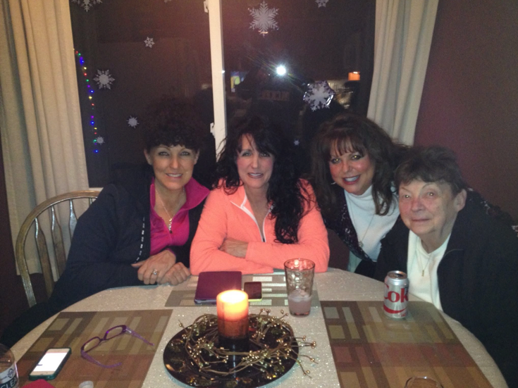 Four women sit around a dining table indoors at a holiday gathering, candles lit and snowflake decorations visible outside the window.
